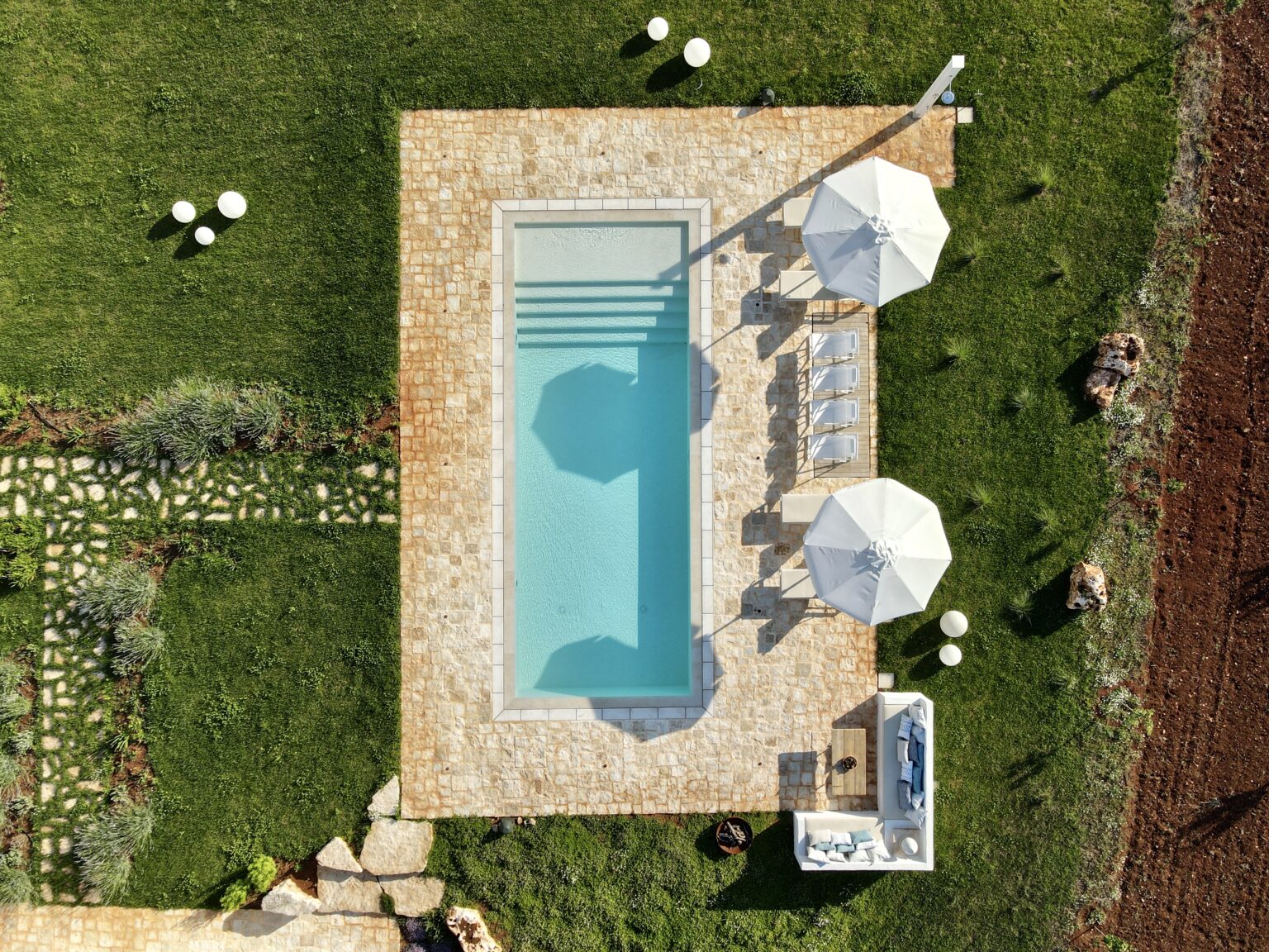 Aerial view of a modern swimming pool surrounded by lush green grass and decorative landscaping, featuring two white umbrellas and sun loungers for relaxation. | Only on SopranoVillas