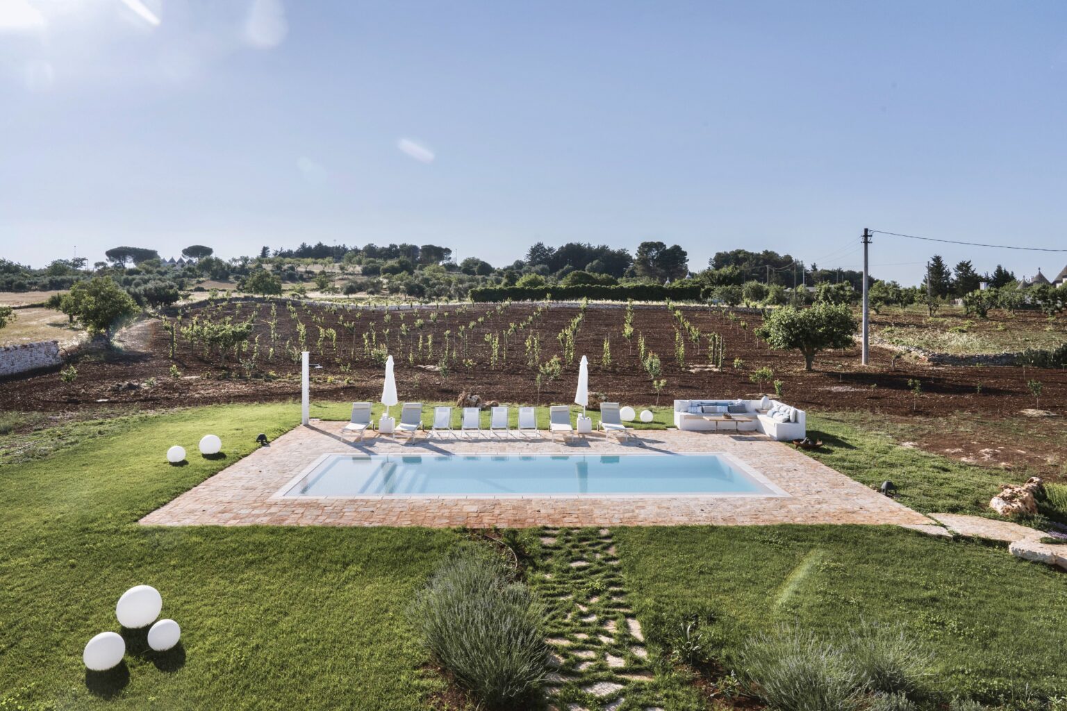 Scenic view of a swimming pool surrounded by lush green grass and modern lounge chairs, with a vineyard in the background under a clear blue sky. Ideal for relaxation and outdoor enjoyment. | Only on SopranoVillas