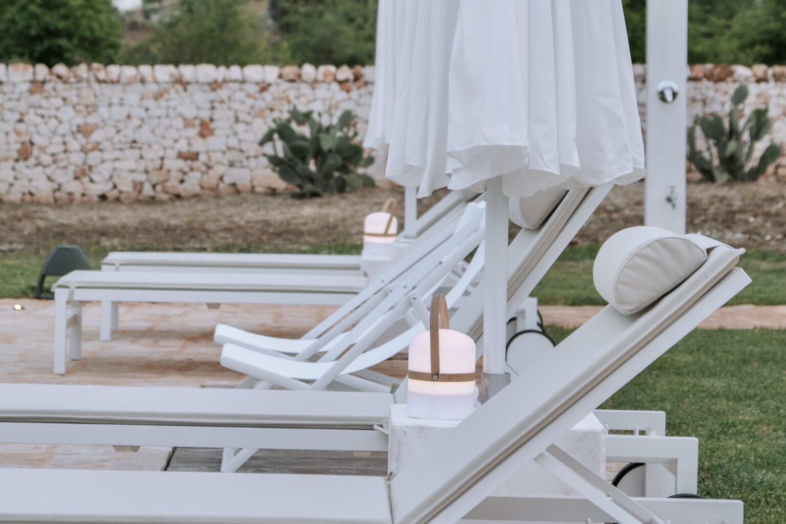 White lounge chairs with umbrellas in a serene outdoor setting, surrounded by greenery and a stone wall, ideal for relaxation and leisure. | Only on SopranoVillas