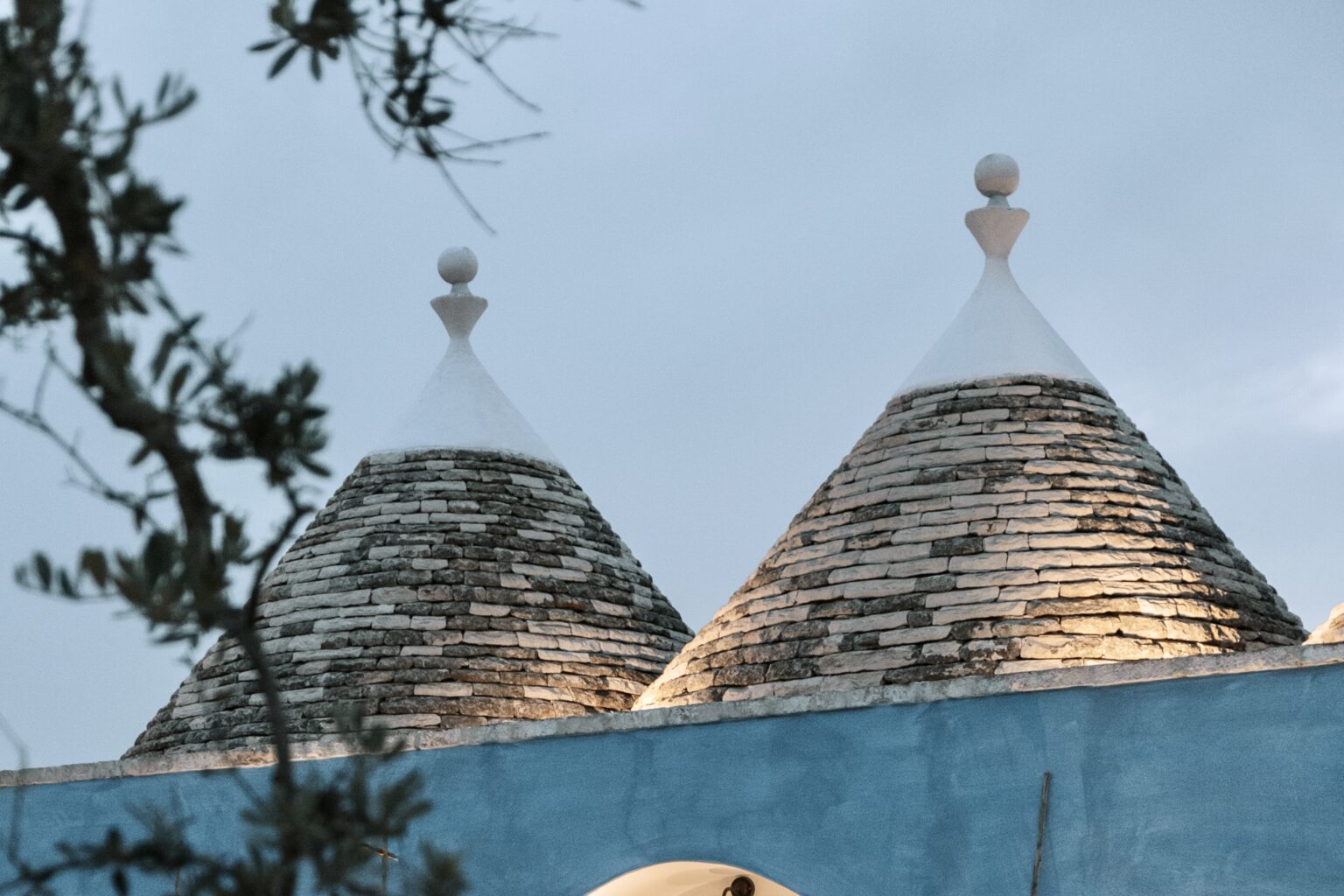 Trulli rooftops with conical stone structures and decorative pinnacles, set against a cloudy sky, showcasing traditional architecture in the Apulia region of Italy. | Only on SopranoVillas