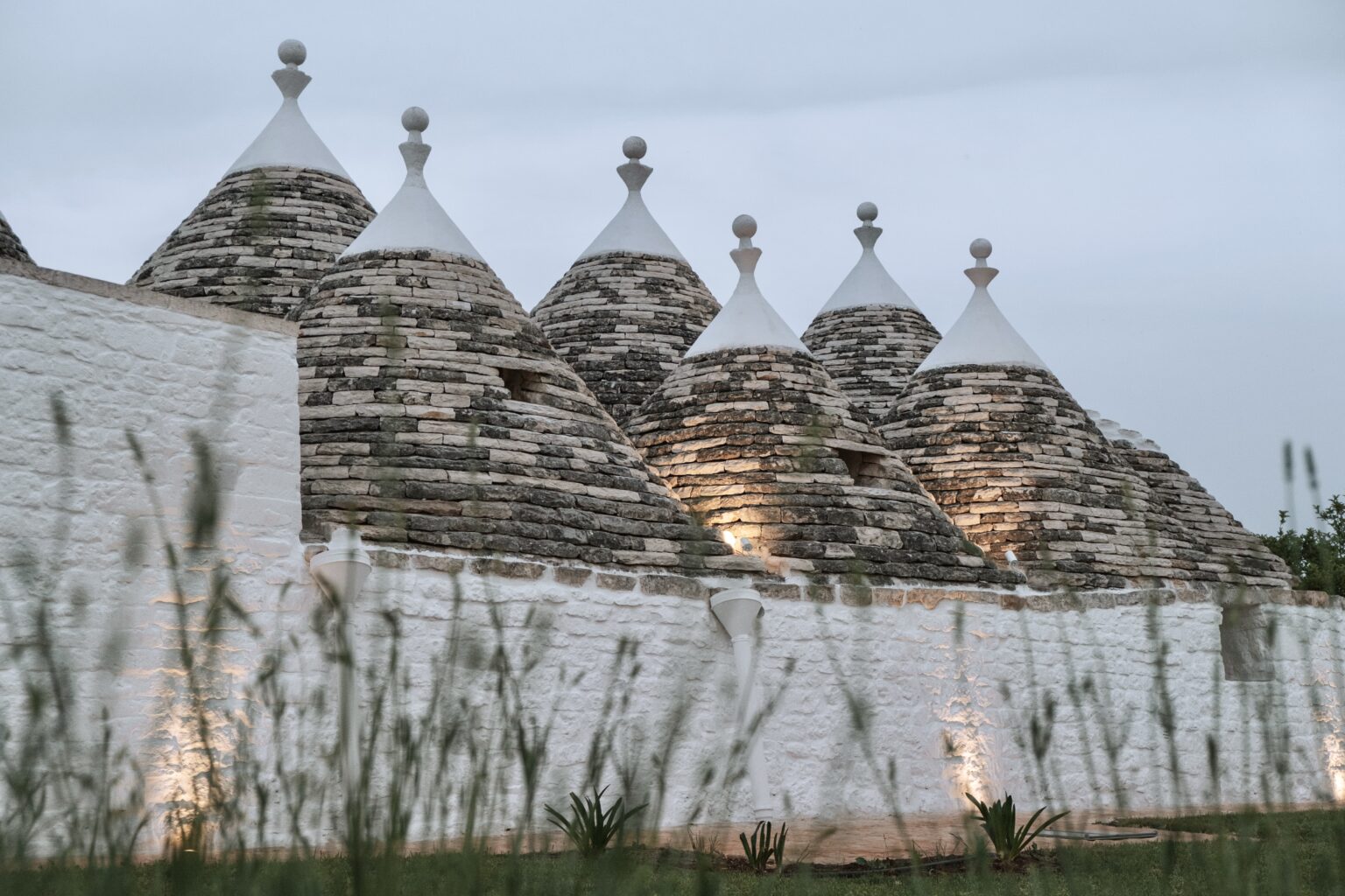 Traditional trulli houses with conical roofs in Puglia, Italy, illuminated at dusk, surrounded by tall grass. | Only on SopranoVillas