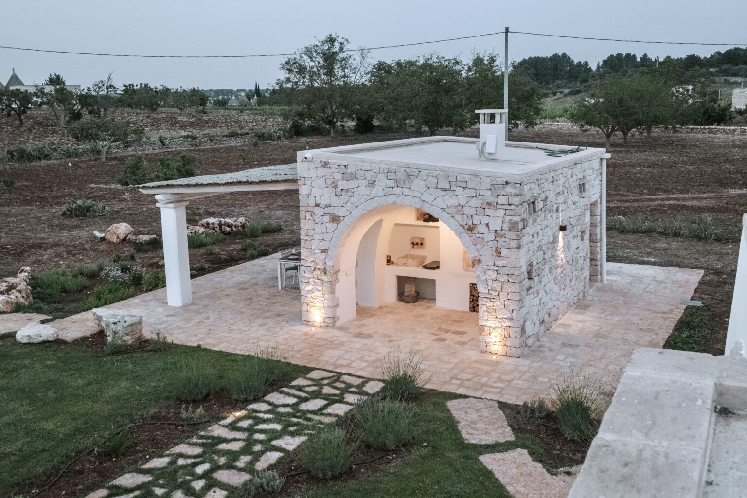 Small stone cottage with an arched entrance, surrounded by landscaped pathways and greenery, set in a tranquil rural landscape during twilight. | Only on SopranoVillas