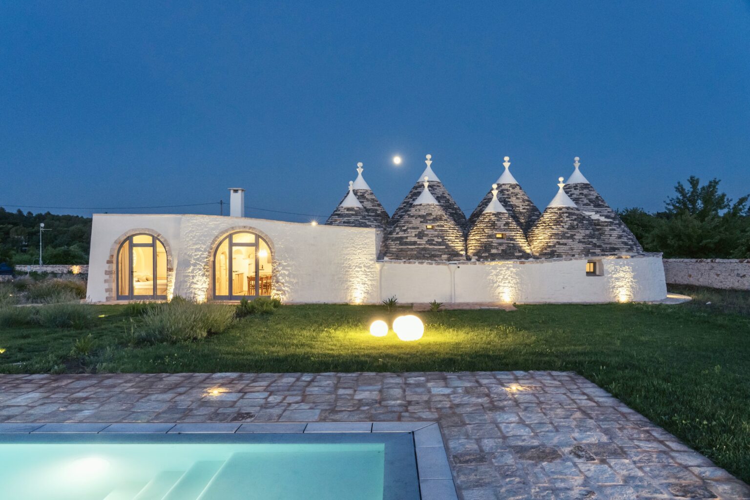 Trulli-style vacation home with conical roofs illuminated at night, surrounded by lush greenery and a swimming pool in the foreground. | Only on SopranoVillas