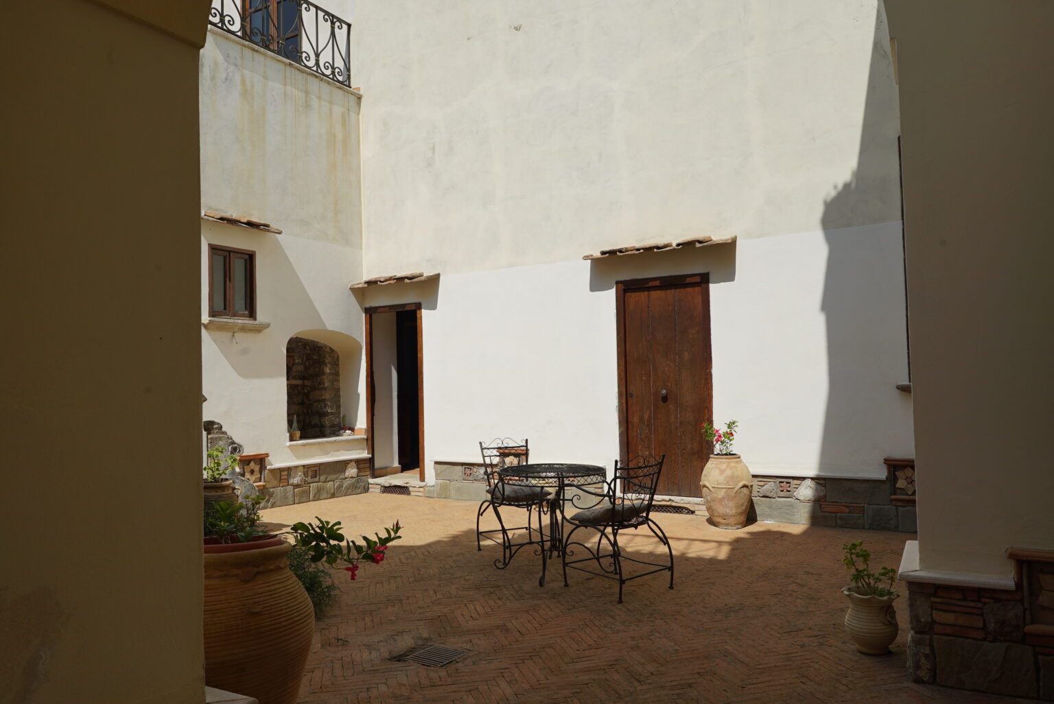 Charming courtyard featuring a round metal table with chairs, surrounded by rustic wooden doors and potted plants, highlighting a serene outdoor space with warm sunlight and textured brick flooring. | Only on SopranoVillas