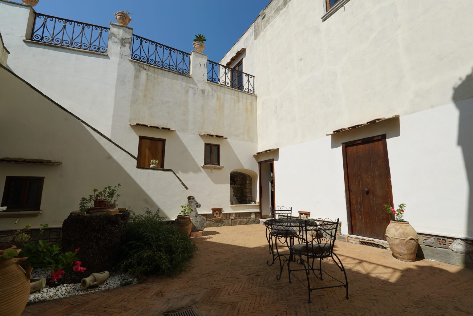Charming courtyard with a rustic design featuring a wrought iron table and chairs, terracotta pots, and a stone wall, illuminated by sunlight under a clear blue sky. | Only on SopranoVillas