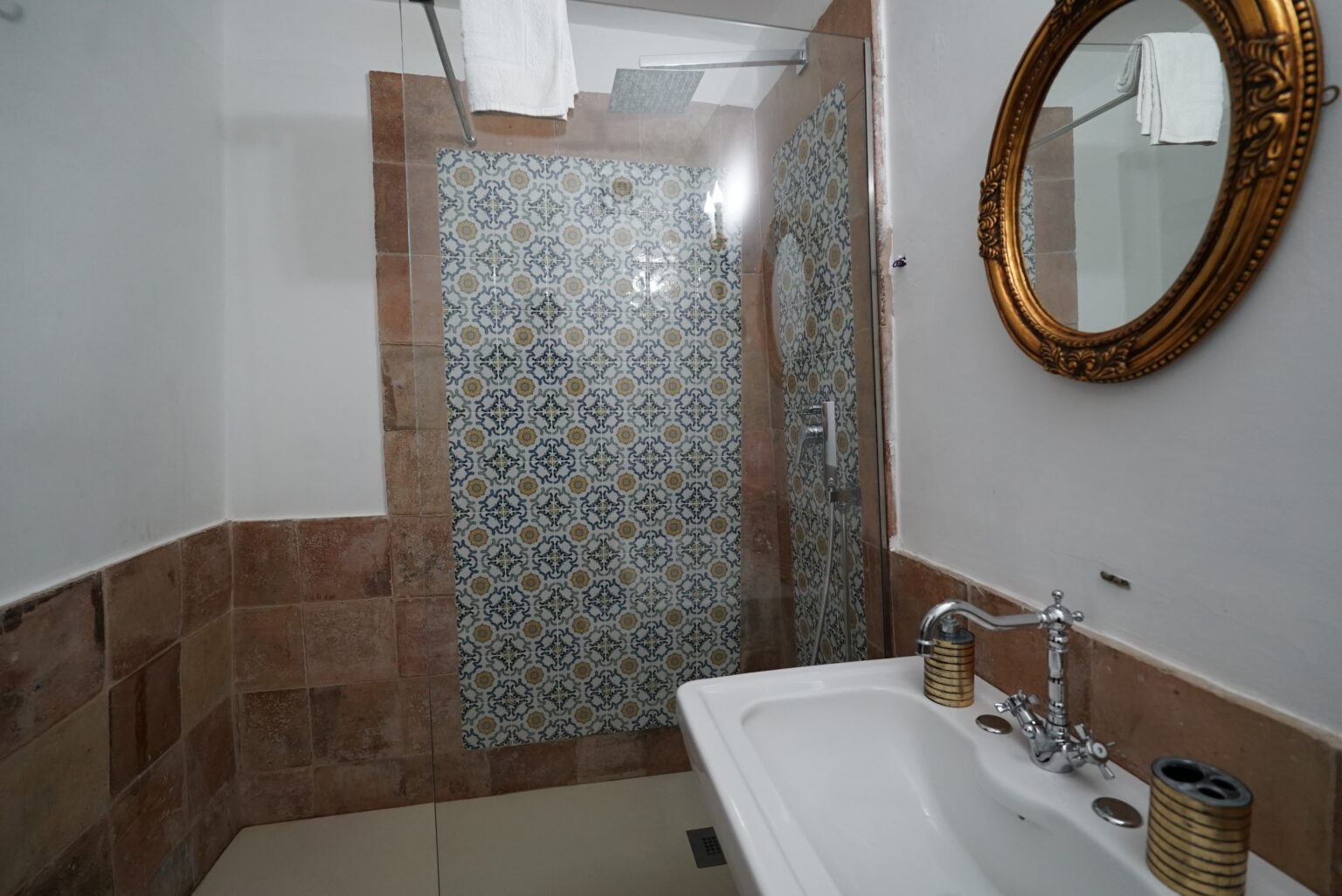 Modern bathroom interior featuring a glass shower enclosure with decorative tiles, a stylish sink with faucets, and an elegant round mirror framed in gold. | Only on SopranoVillas