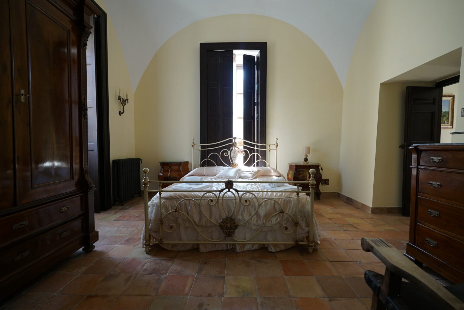 Cozy bedroom featuring a wrought iron bed with light bedding, wooden furniture, and a large window allowing natural light to illuminate the space, set in a rustic interior with terracotta flooring. | Only on SopranoVillas