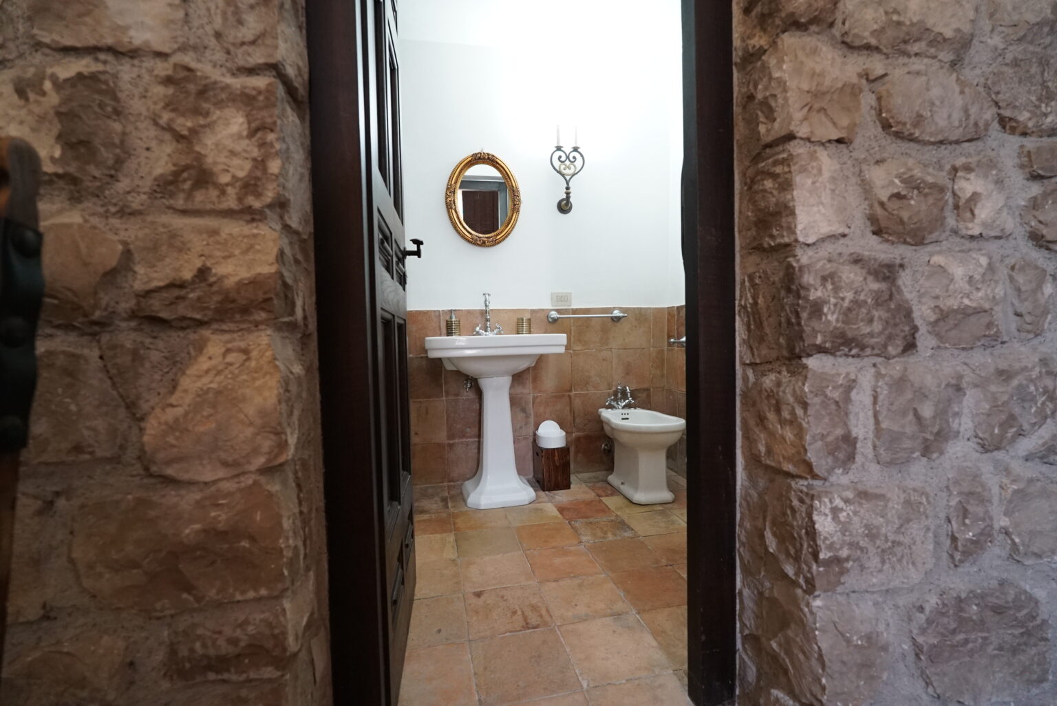 View of a rustic bathroom featuring stone walls, a pedestal sink, a toilet, and a round mirror, captured from the doorway. The warm tones of the tile floor complement the natural materials. | Only on SopranoVillas