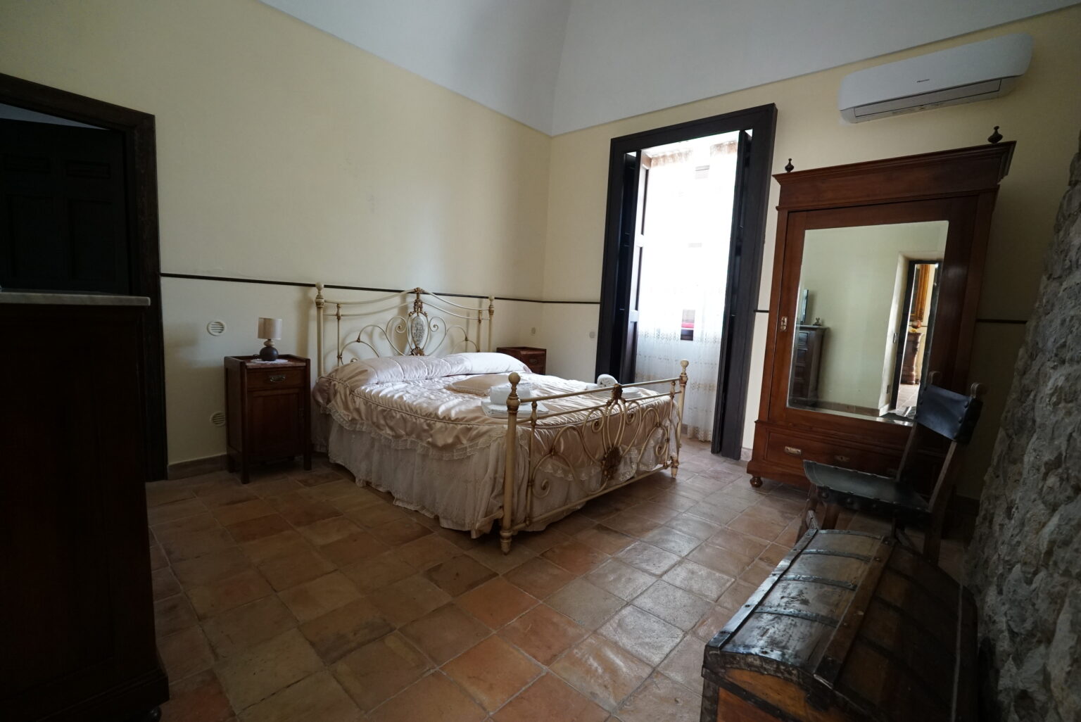 Cozy bedroom featuring a vintage metal bed with a decorative headboard, wooden furniture including a dresser and a chair, and large windows allowing natural light. The room has tiled flooring and a warm color palette, creating an inviting atmosphere. | Only on SopranoVillas