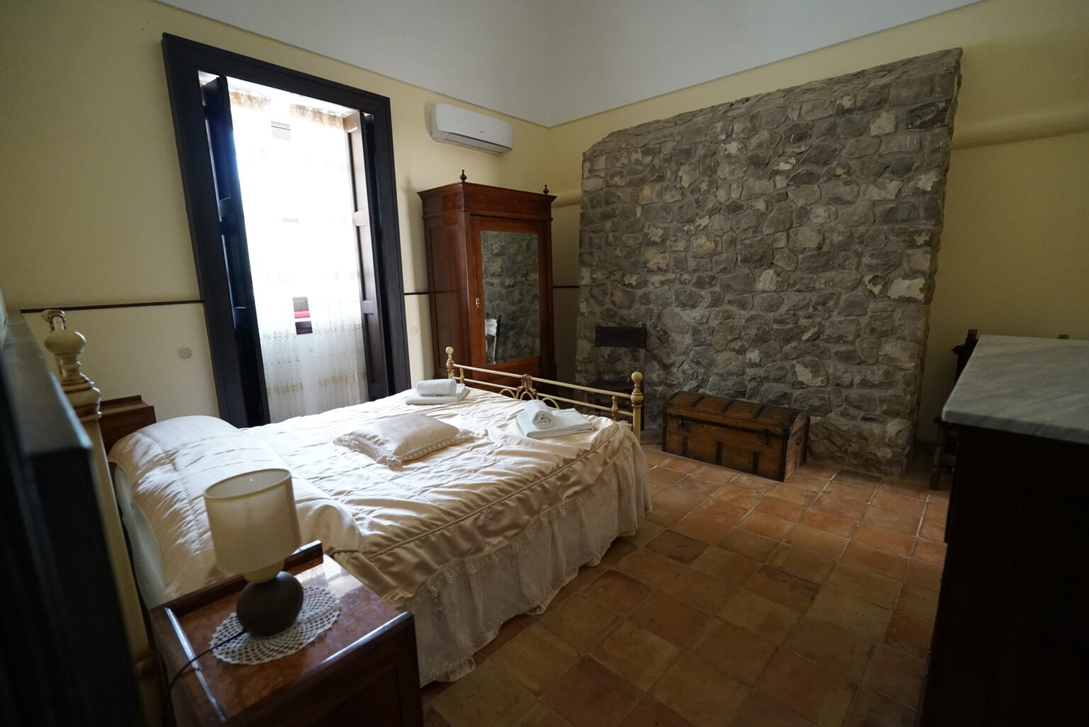 Cozy bedroom featuring a brass bed with white linens, a wooden wardrobe, and a stone wall. The room has a large window with sheer curtains, allowing natural light to brighten the space. A vintage wooden trunk and a bedside lamp add charm to the decor. | Only on SopranoVillas