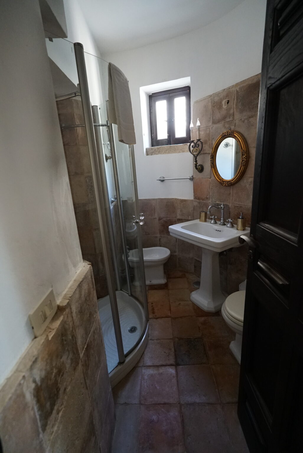 Modern bathroom featuring a glass shower, pedestal sink, and decorative mirror, adorned with natural stone tiles and warm lighting. | Only on SopranoVillas