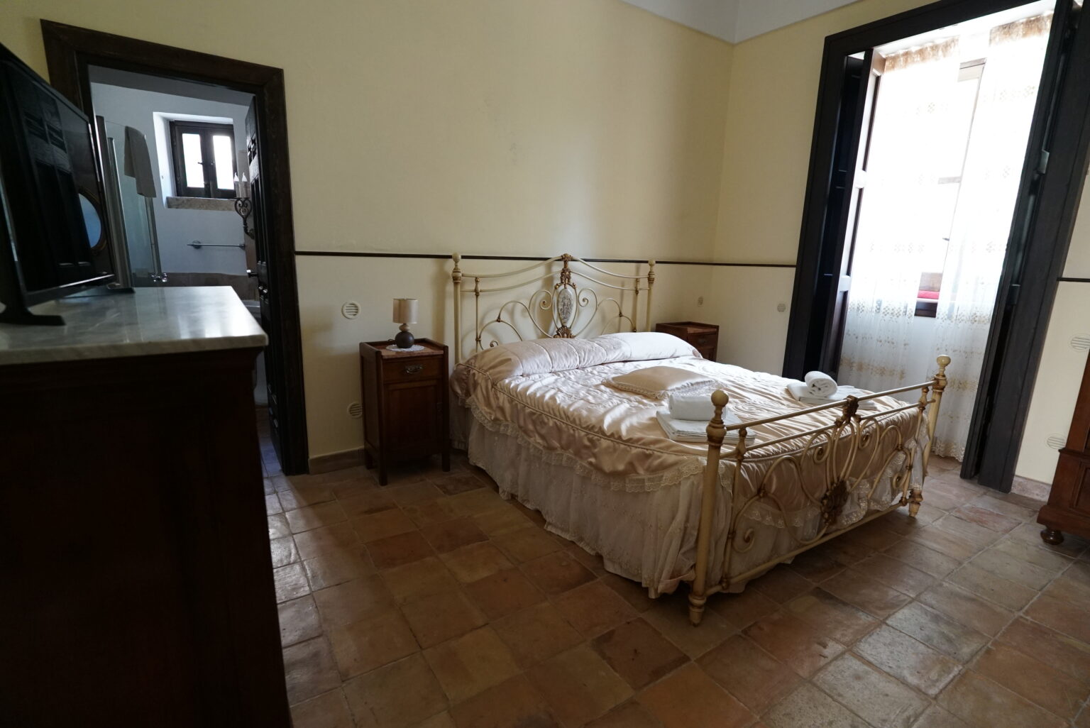 Cozy bedroom featuring a vintage iron bed with soft bedding, two wooden nightstands, and a window draped with sheer curtains. The room has tiled flooring and a warm yellow color scheme, creating a welcoming atmosphere. A doorway leads to a bathroom area, enhancing the room's functionality. | Only on SopranoVillas