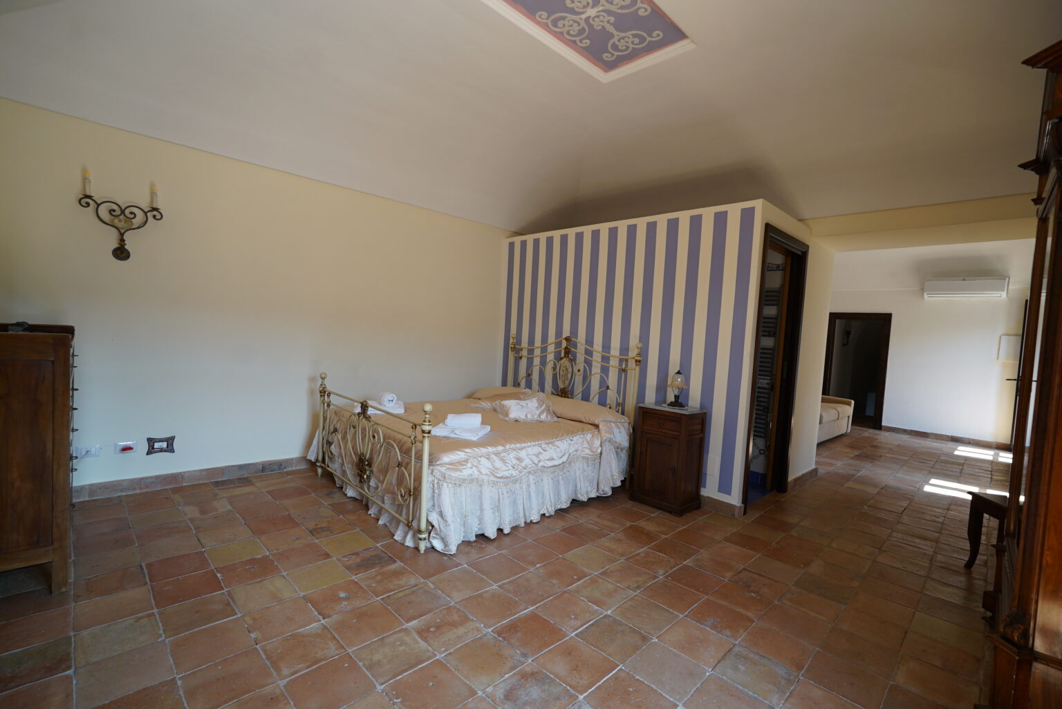 Spacious bedroom featuring a double bed with decorative linens, terracotta tile flooring, and striped wallpaper, complemented by vintage furniture and natural light. | Only on SopranoVillas