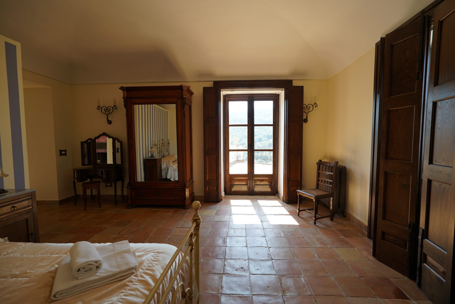 Spacious bedroom with wooden furniture, large windows allowing natural light, and a rustic tile floor, featuring a bed, a dresser, and a chair. | Only on SopranoVillas