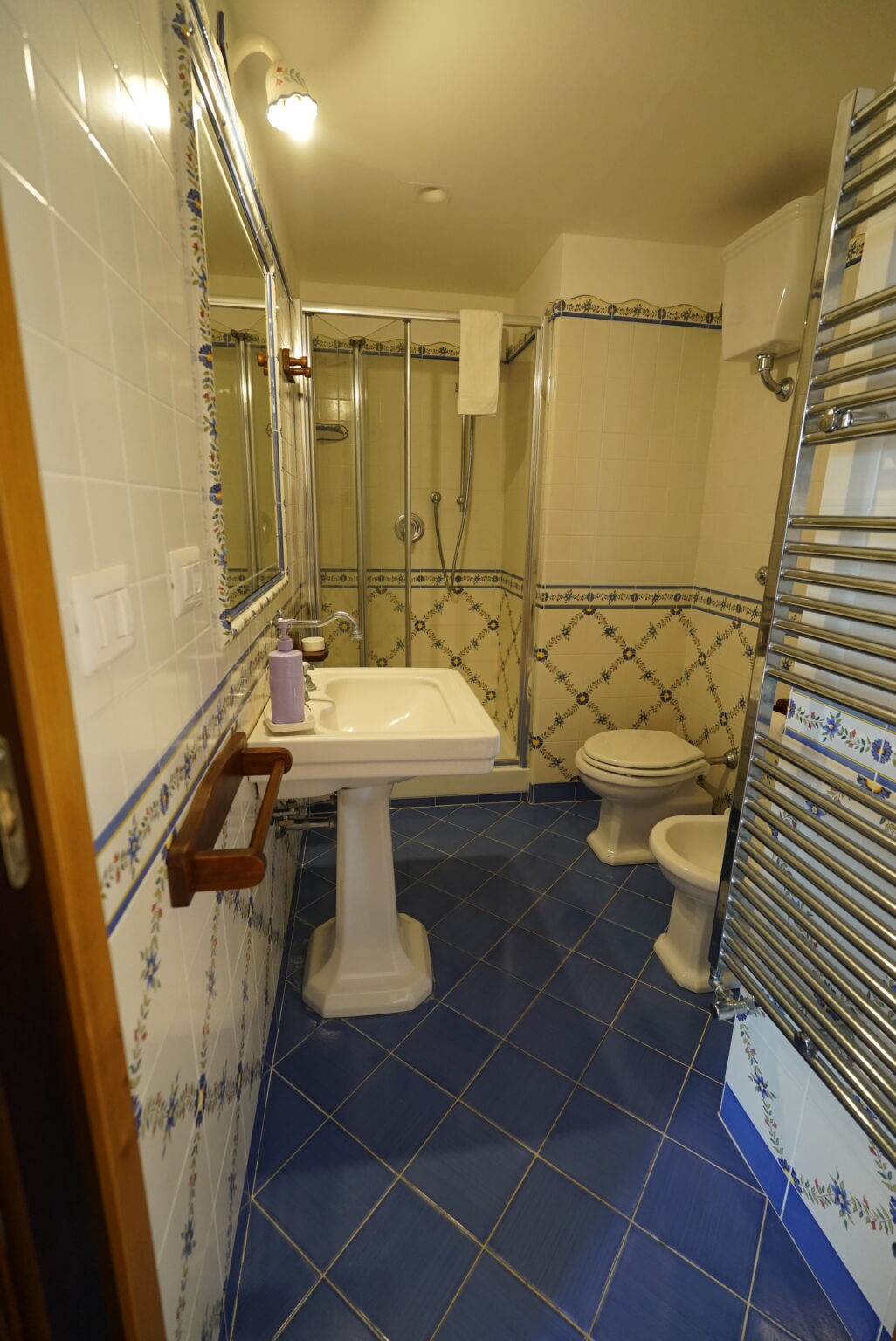 Modern bathroom featuring a glass shower enclosure, white pedestal sink, and toilet. The space is decorated with blue and floral patterned tiles, complemented by a heated towel rack. Ample lighting and a mirror enhance the bright and clean ambiance. | Only on SopranoVillas