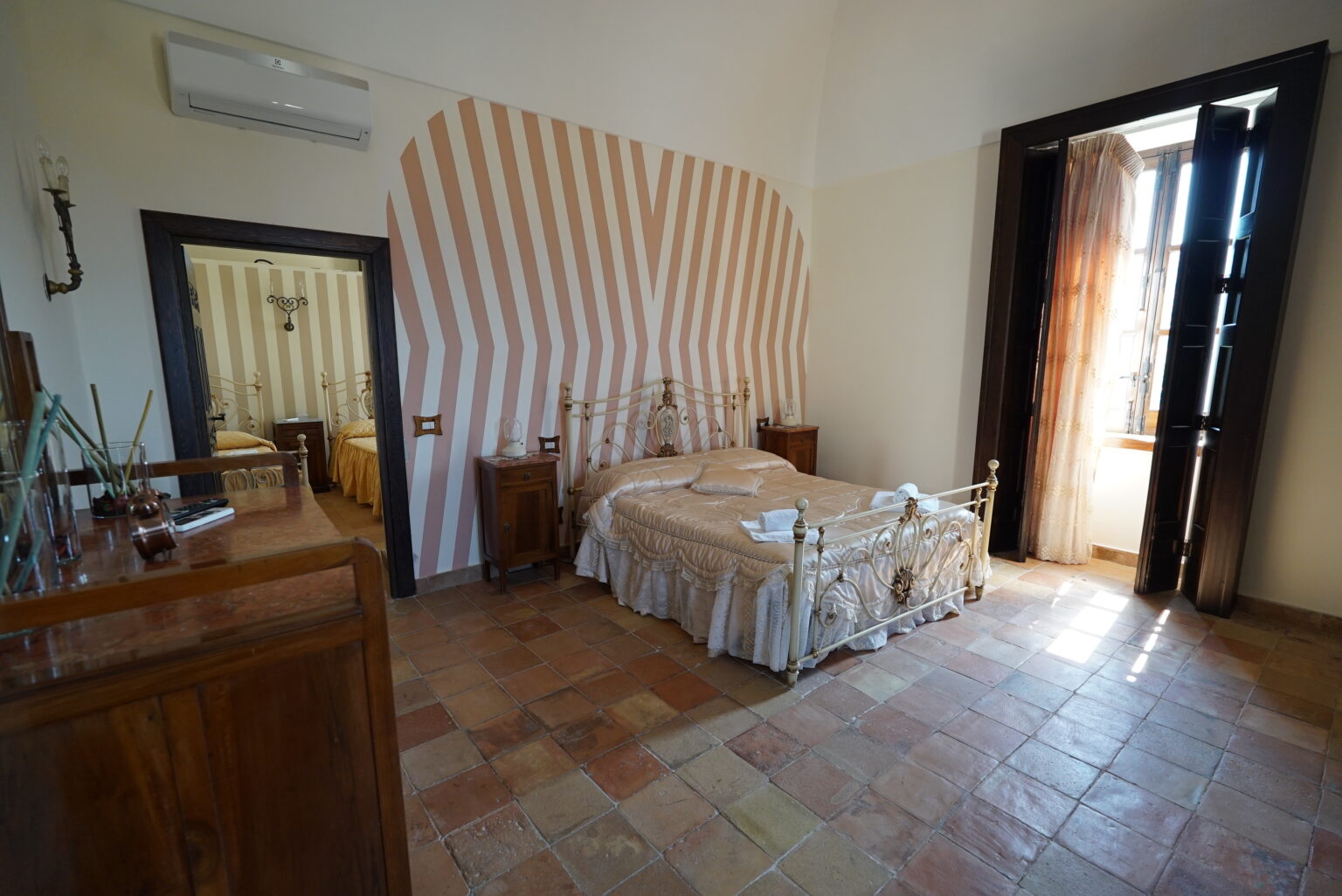 Spacious and inviting bedroom featuring a vintage metal bed, elegant decor, and large windows allowing natural light. The room showcases a unique striped wall design and tiled flooring, creating a warm and cozy atmosphere. | Only on SopranoVillas