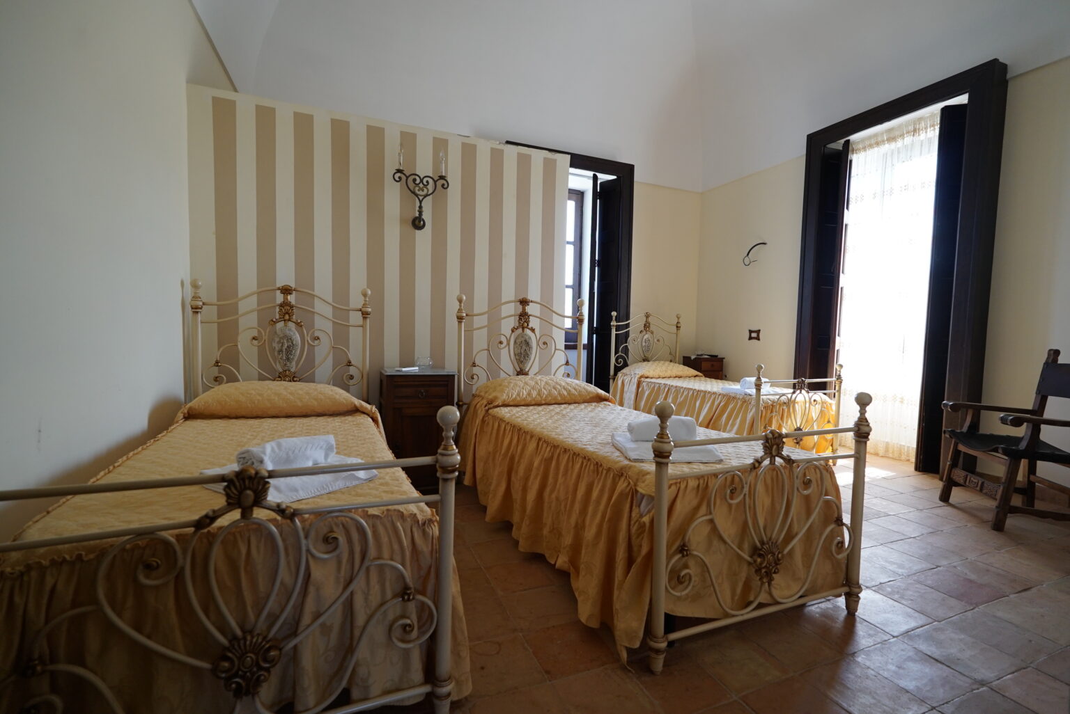 Cozy guest room featuring three twin beds with yellow bedding, vintage metal bed frames, and a bright window with sheer curtains. The room has a warm, inviting atmosphere with striped wallpaper and a wooden chair. Ideal for families or groups seeking comfortable accommodation. | Only on SopranoVillas