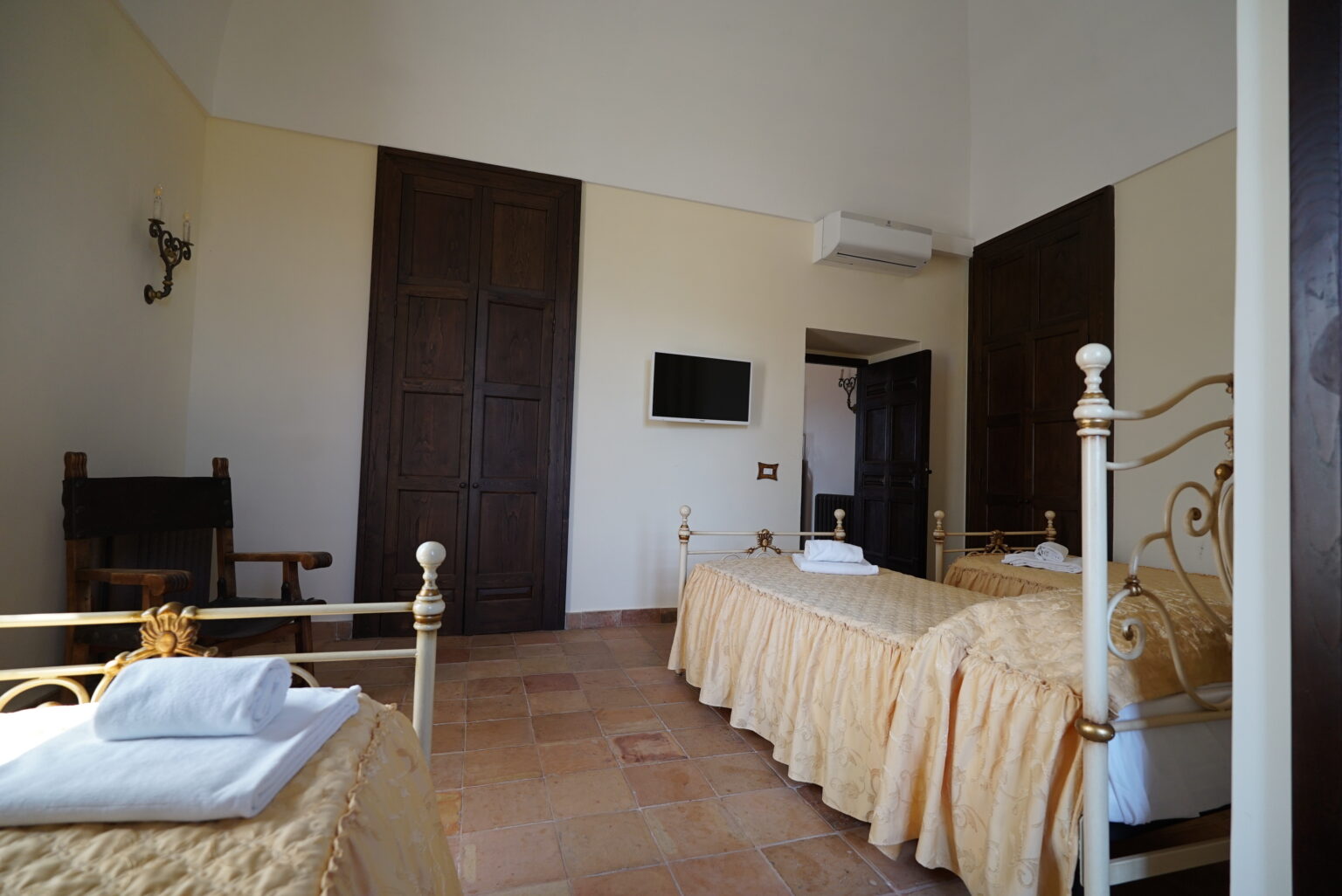 Cozy hotel room featuring two twin beds with elegant gold bedding, a flat-screen TV mounted on the wall, and wooden doors. The space is well-lit with natural light and includes a small seating area, perfect for relaxation. | Only on SopranoVillas