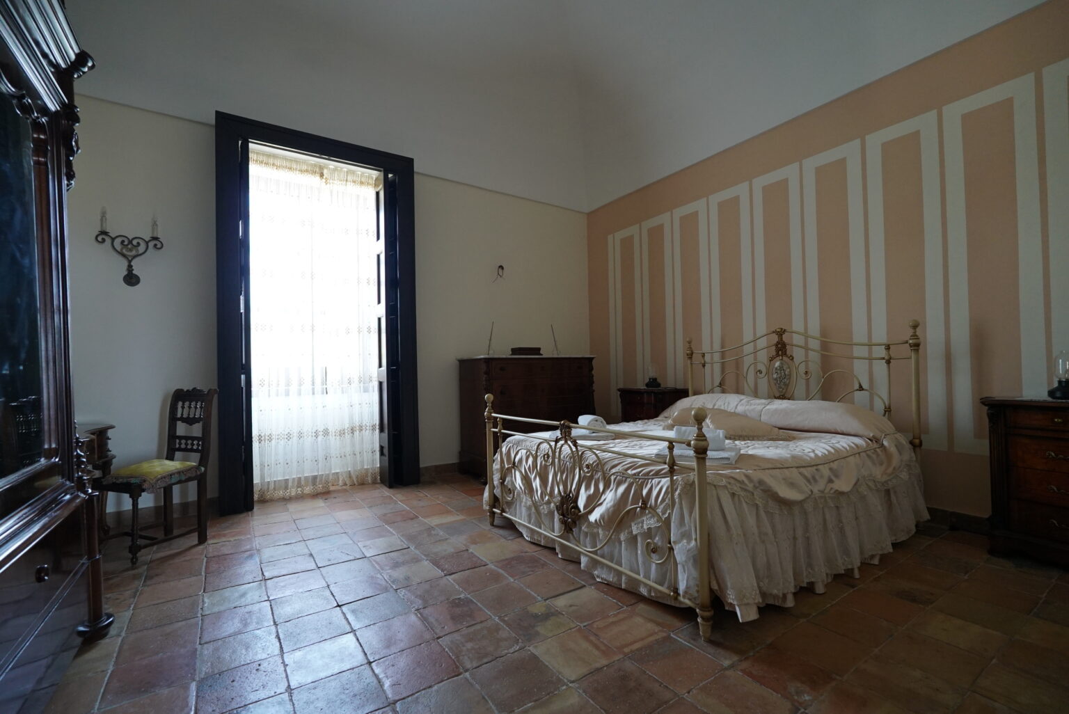 Cozy bedroom interior featuring a vintage bed with soft bedding, a wooden dresser, and a large window allowing natural light to enter, complemented by elegant wall decor and tiled flooring. | Only on SopranoVillas