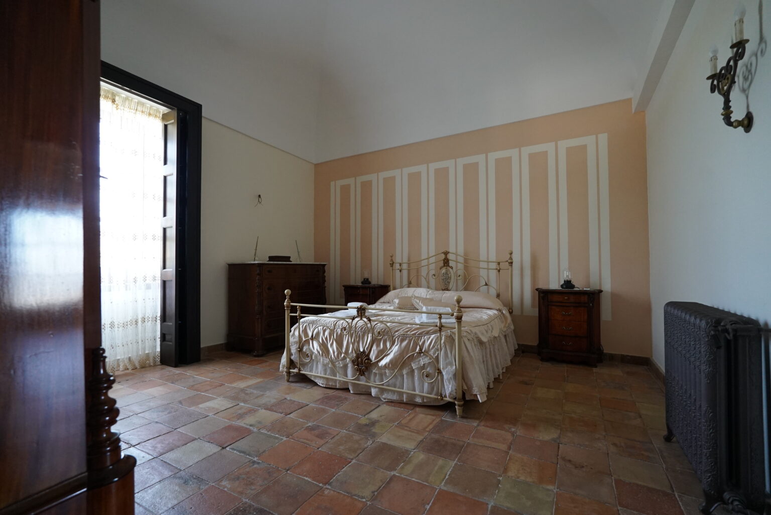 Spacious bedroom featuring a large bed with a decorative cover, wooden furniture, and natural light streaming through a window, showcasing a warm and inviting atmosphere. | Only on SopranoVillas