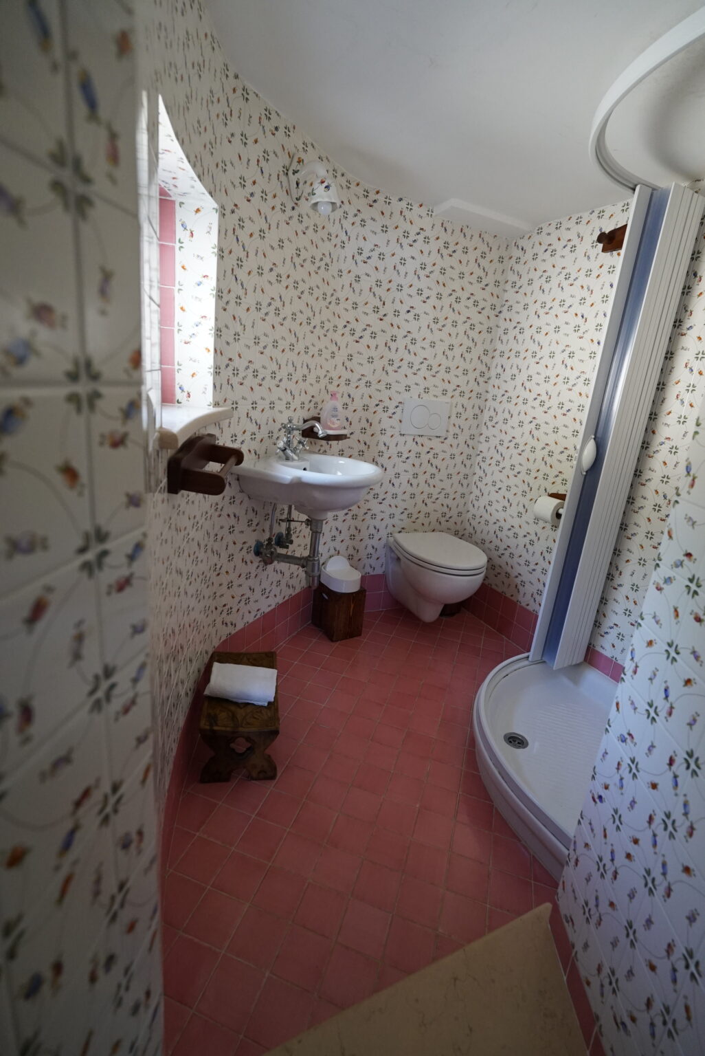Cozy bathroom featuring floral wallpaper, a small sink, toilet, and shower, with pink flooring and natural light. | Only on SopranoVillas