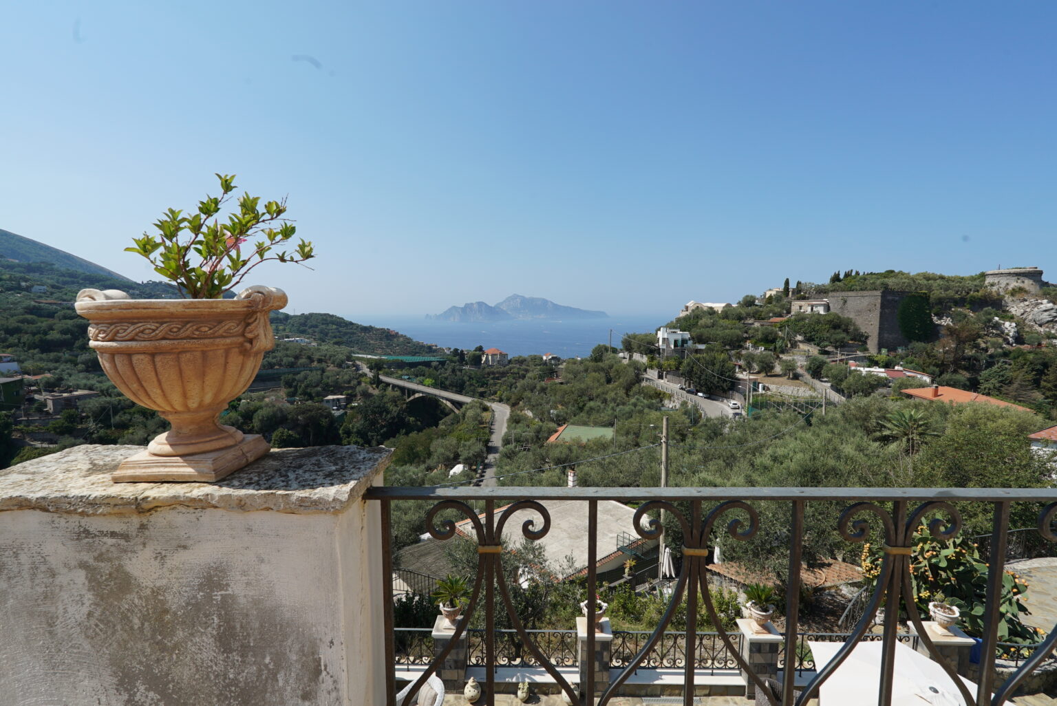 Scenic view from a balcony featuring a decorative flower pot, overlooking lush green hills and distant mountains under a clear blue sky. | Only on SopranoVillas