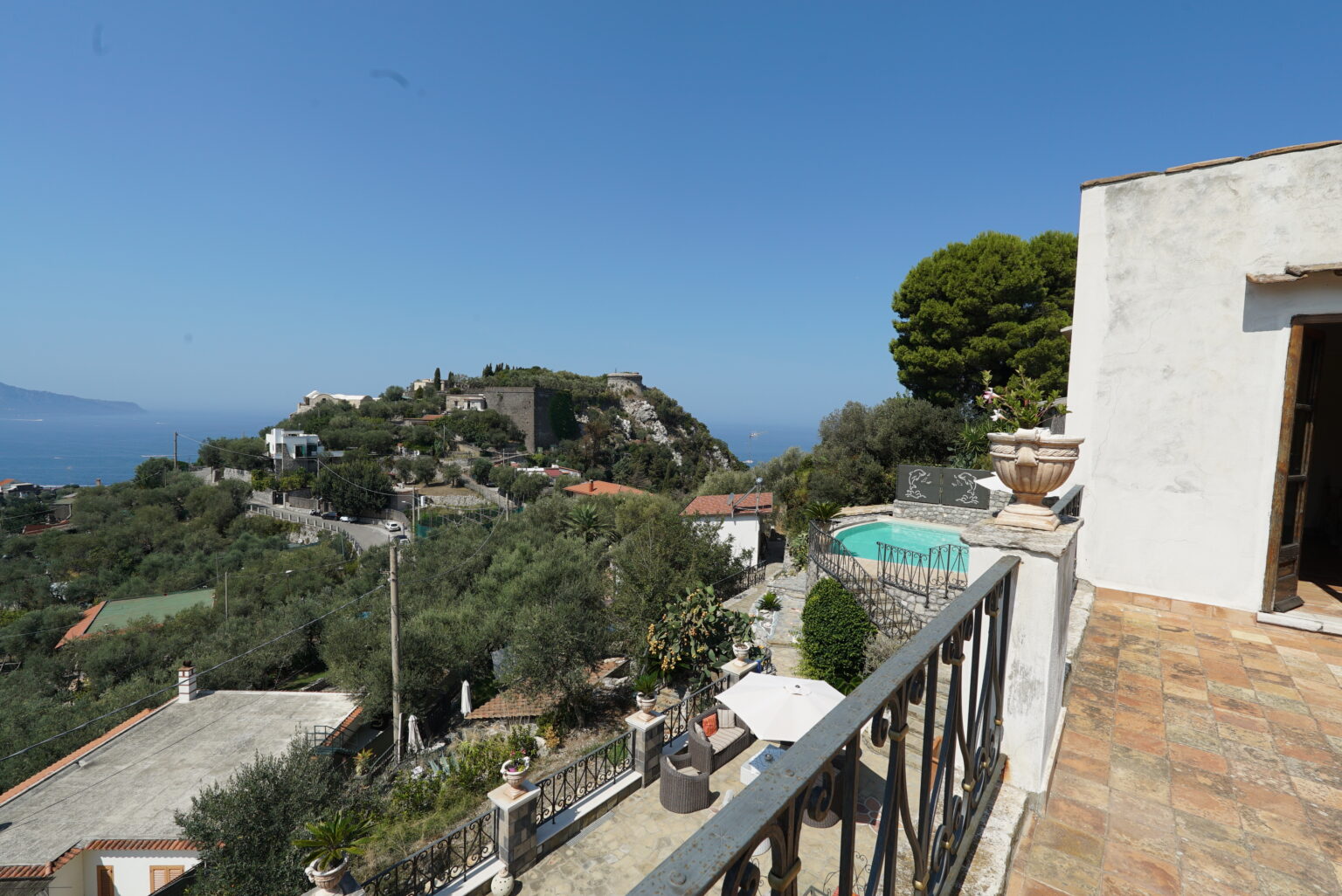 Scenic view from a balcony overlooking a hillside villa with a swimming pool, surrounded by lush greenery and clear blue skies. | Only on SopranoVillas