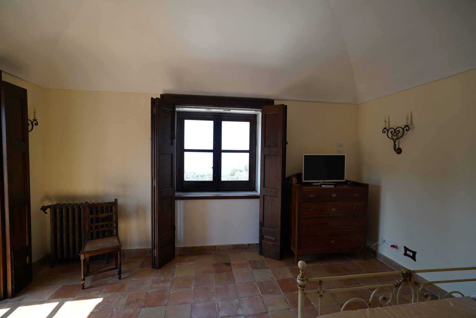 Cozy interior of a bedroom featuring a wooden chair, a window with wooden shutters, a small television on a dresser, and warm-toned tiled flooring. | Only on SopranoVillas