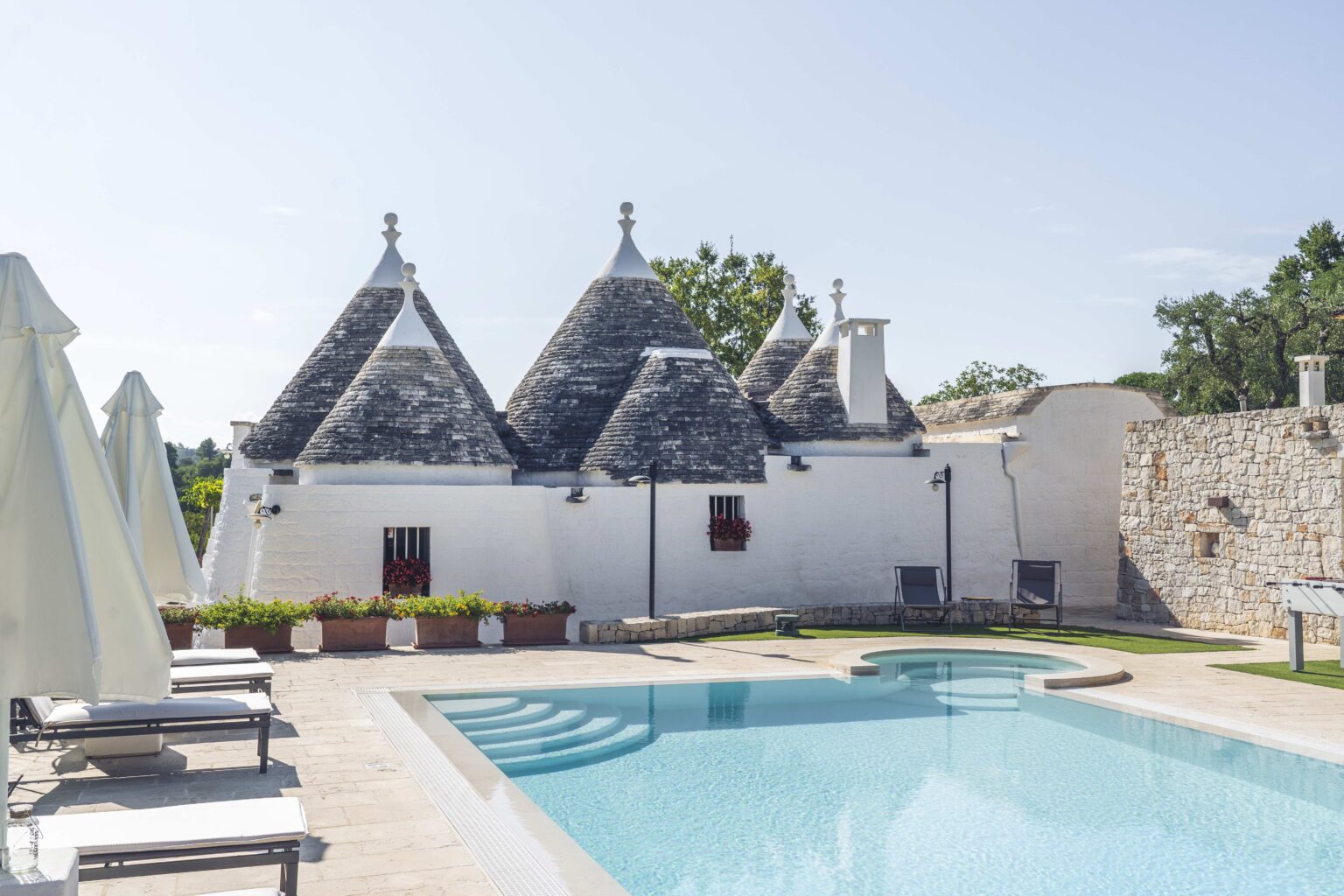 Tranquil pool area with traditional trullo houses in Puglia, Italy, showcasing unique stone architecture and lush greenery under a clear blue sky. | Only on SopranoVillas