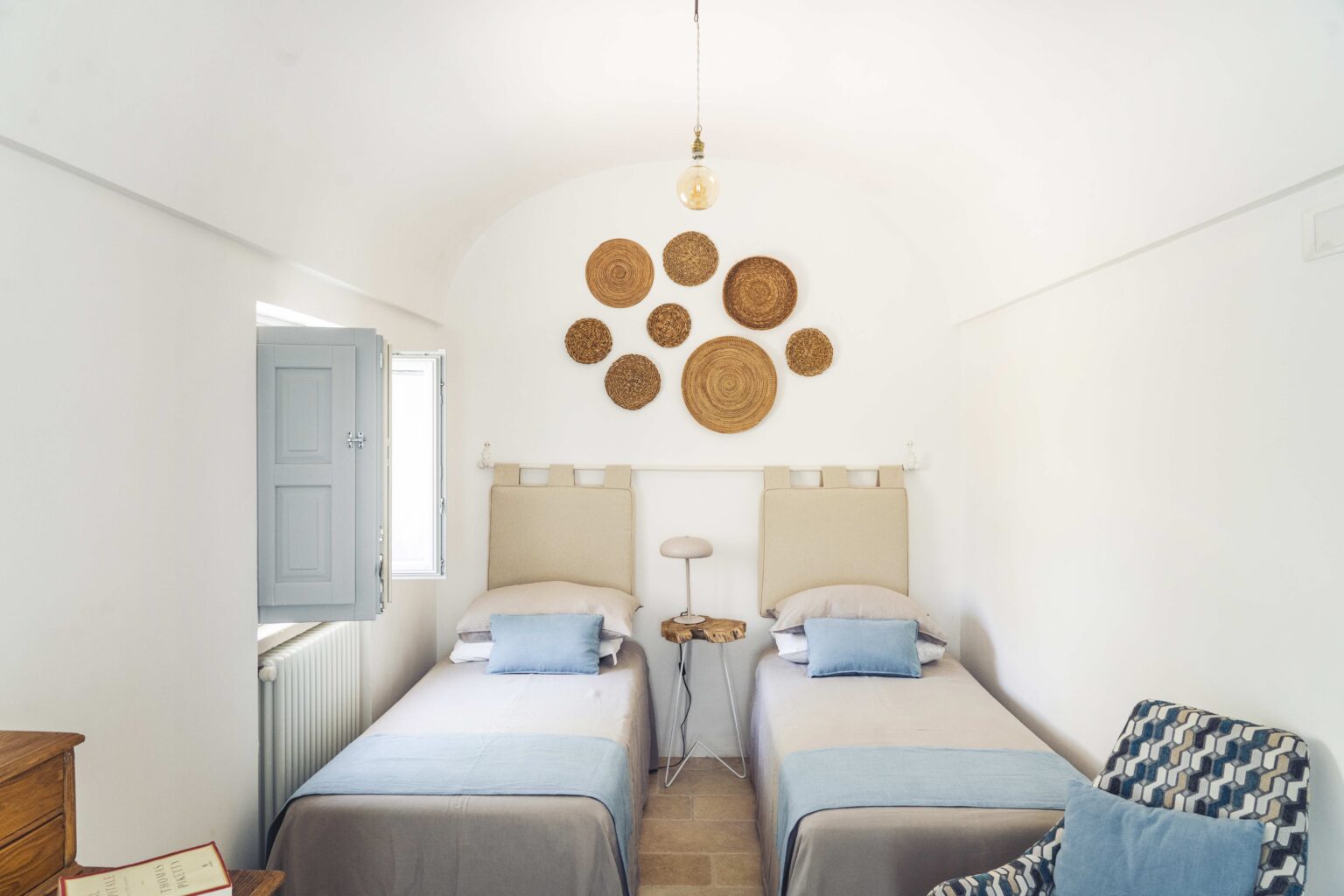 Bright and airy bedroom featuring two twin beds with beige and blue bedding, a wooden side table with a modern lamp, and decorative woven wall art. Natural light streams in through an open window, enhancing the minimalist decor. Ideal for a relaxing getaway. | Only on SopranoVillas