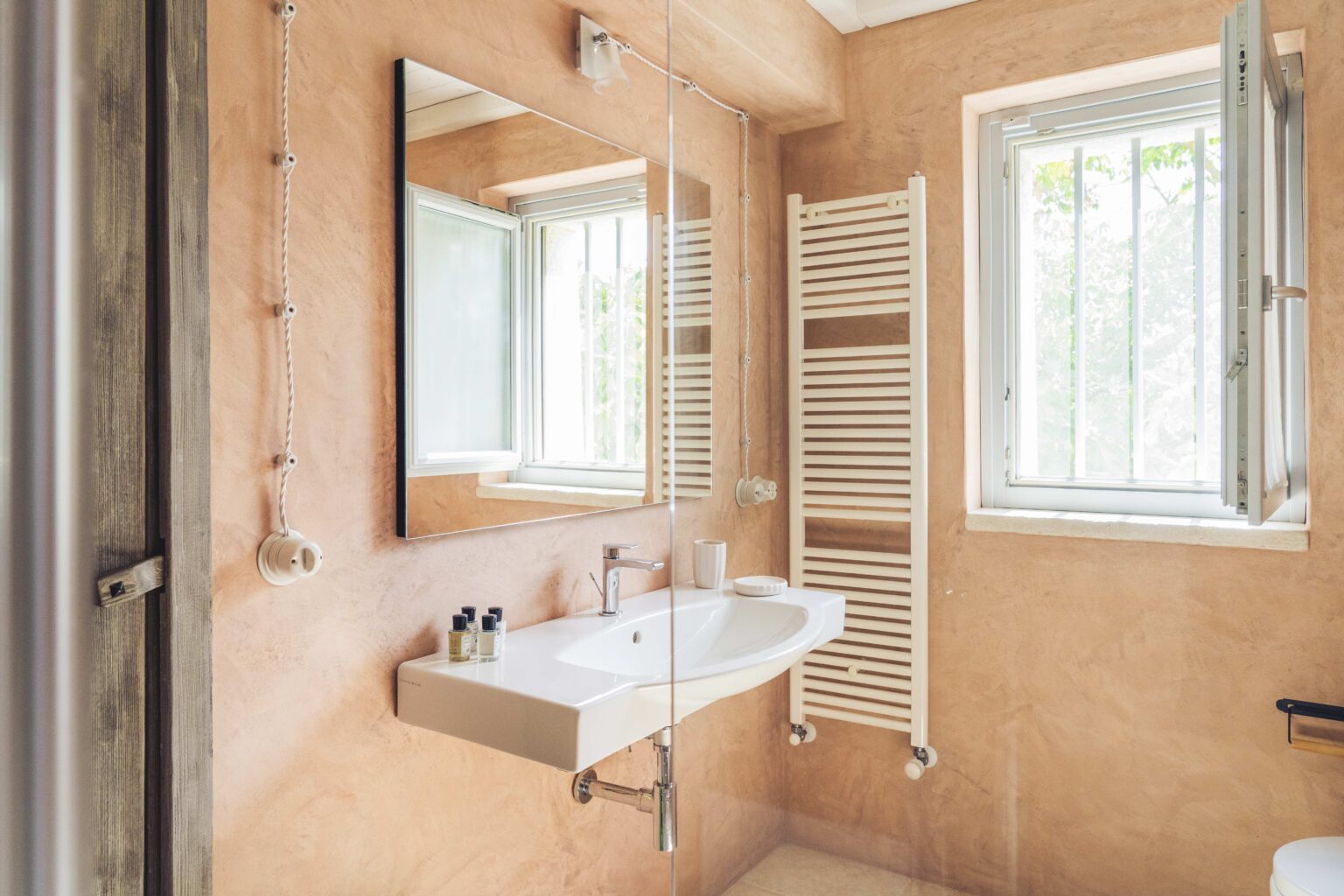 Modern bathroom with peach-colored walls, a large mirror, a sleek sink, and a towel radiator, featuring natural light from a window. | Only on SopranoVillas