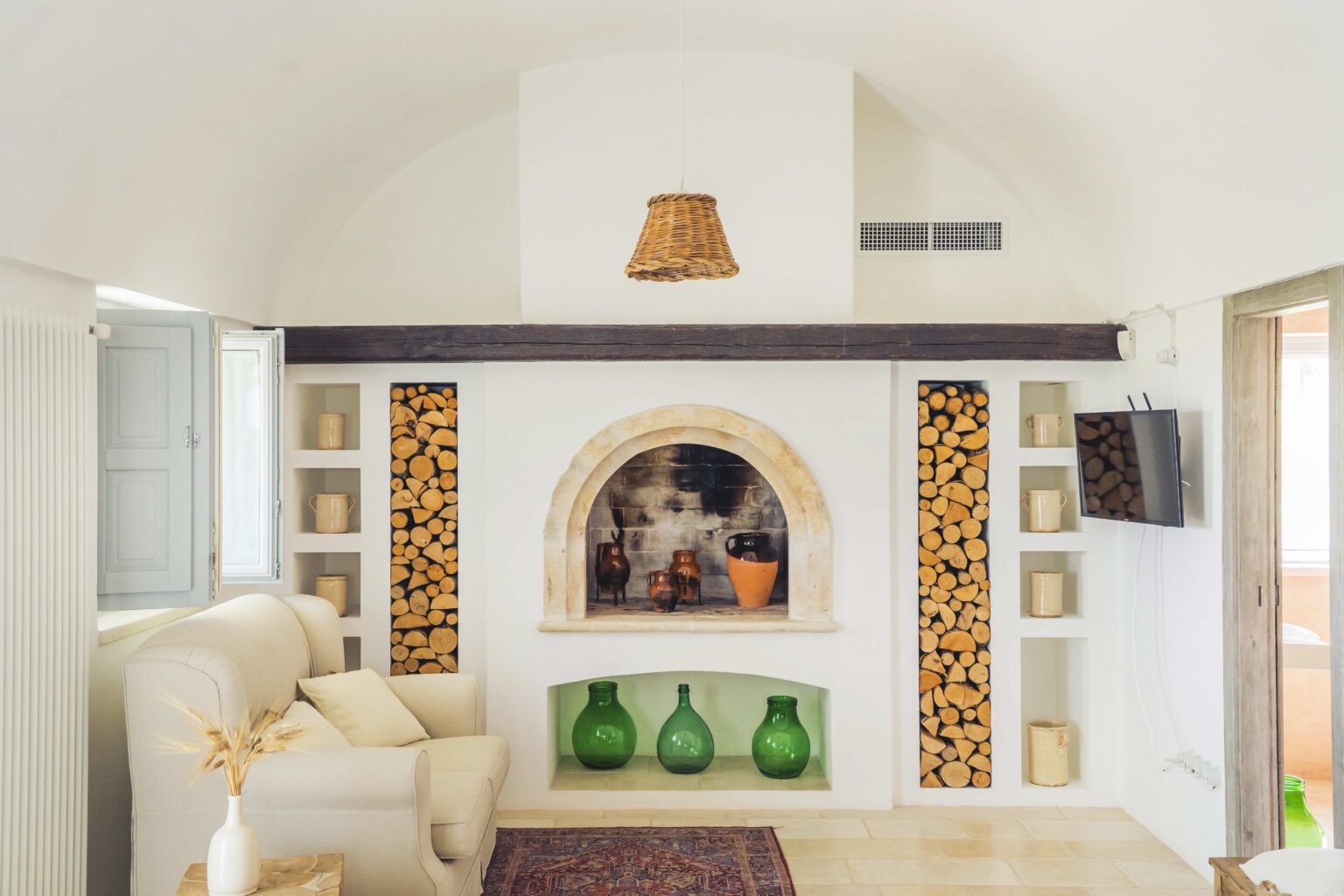 Cozy living room featuring a white sofa, a decorative fireplace with stacked logs, green glass vases, and warm lighting from a woven lamp. The minimalist design incorporates natural elements, enhancing the inviting atmosphere. | Only on SopranoVillas