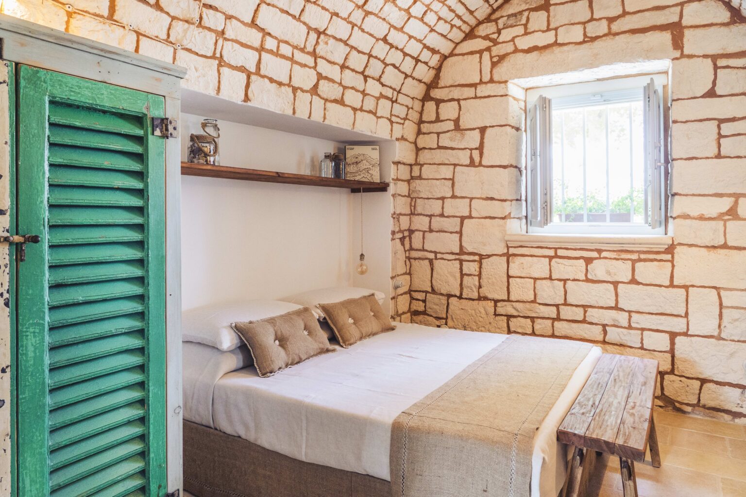 Cozy bedroom featuring a stone wall, a comfortable bed with neutral bedding, decorative cushions, a rustic wooden bench, and a vintage green shutter door, creating a warm and inviting atmosphere. | Only on SopranoVillas