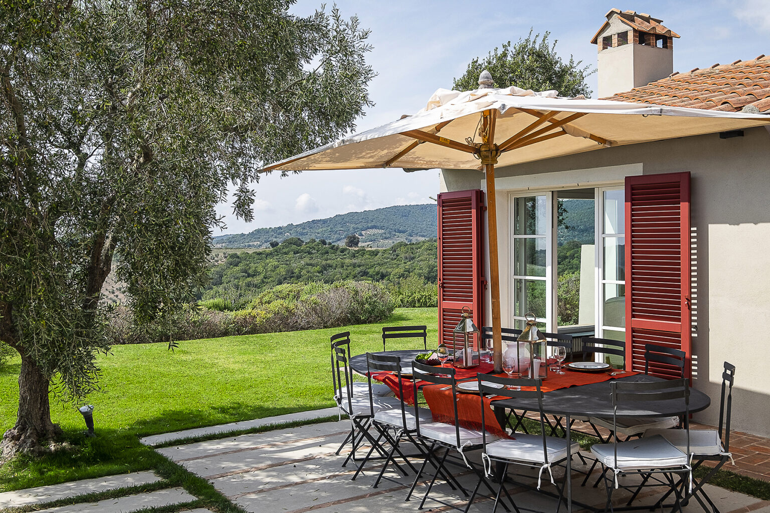 Outdoor dining area with a large umbrella, surrounded by lush greenery and a scenic view, featuring a stylish table set for meals near a house with red shutters. | Only on SopranoVillas