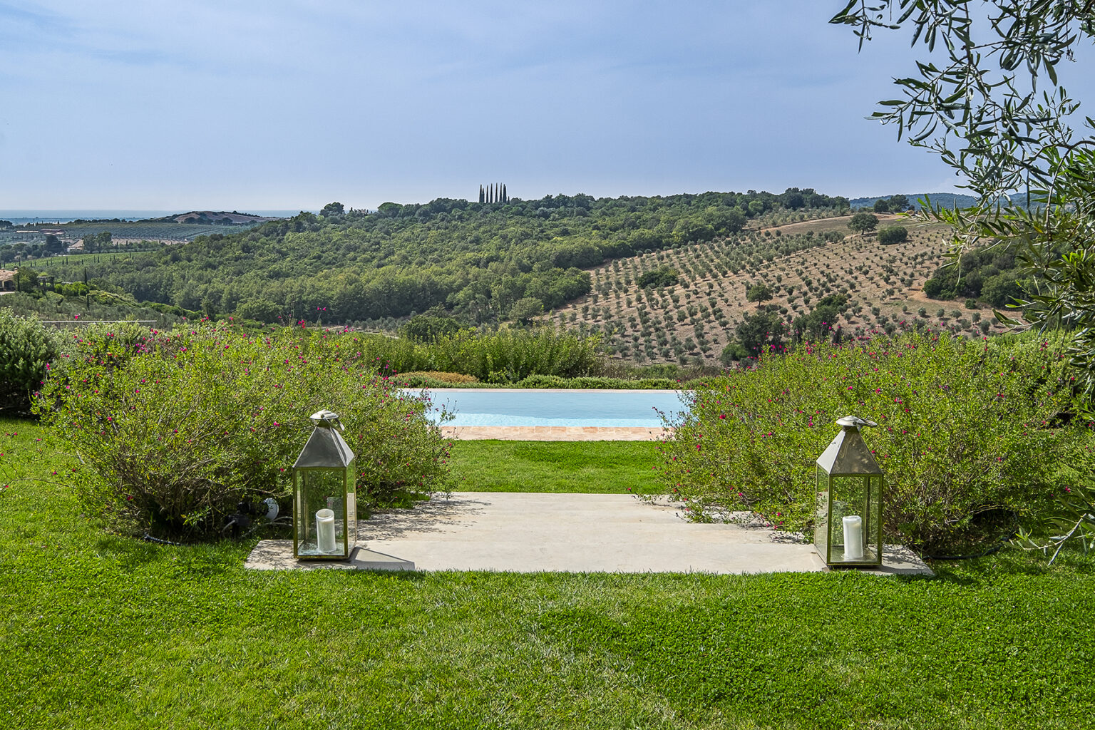 Scenic view of a landscaped garden leading to a swimming pool, surrounded by green hills and vineyards under a clear sky. Two lanterns frame the path, enhancing the tranquil outdoor setting. | Only on SopranoVillas