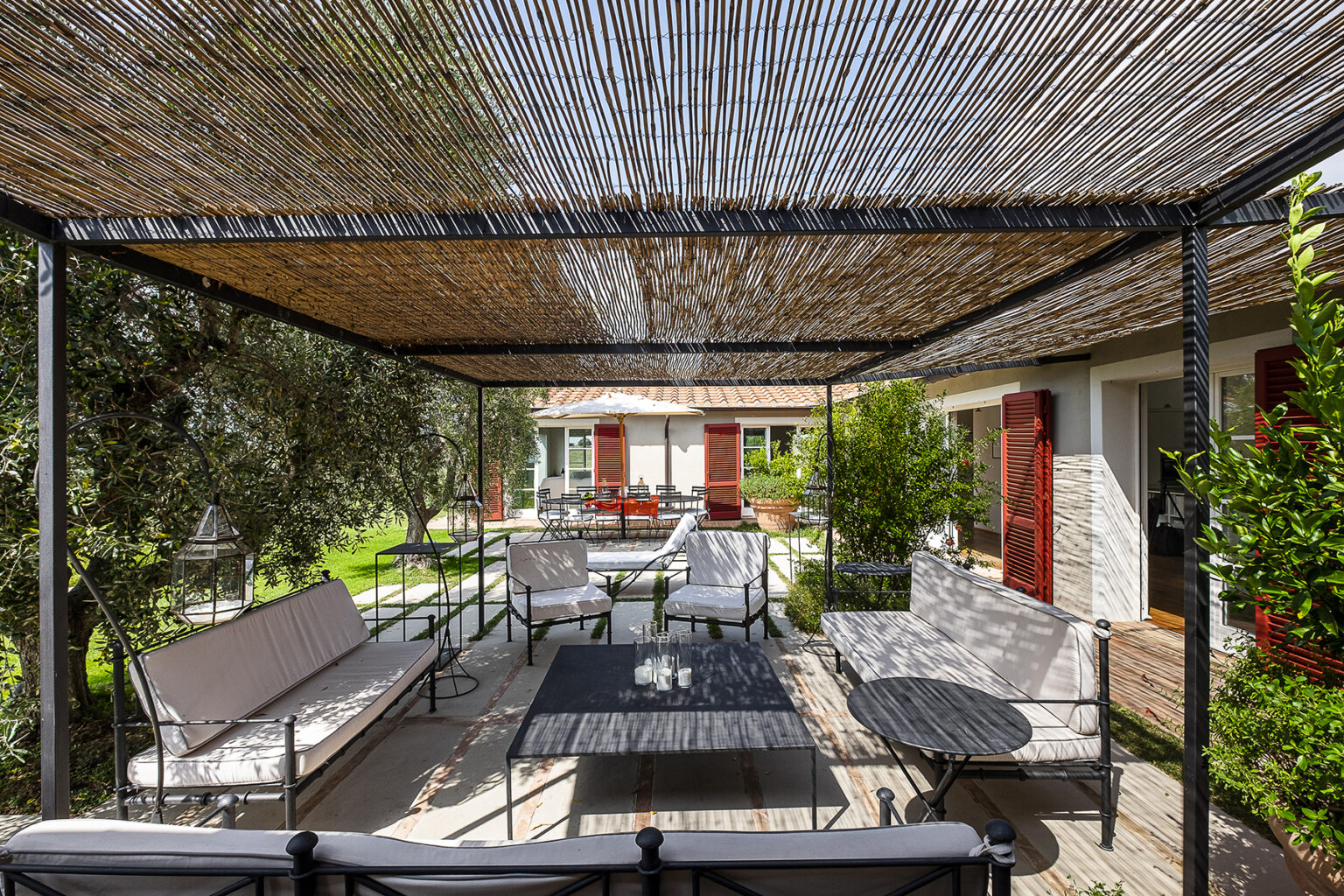 Outdoor patio area with a shaded seating arrangement under a bamboo cover, featuring modern furniture and lush greenery, ideal for relaxation and entertaining. | Only on SopranoVillas