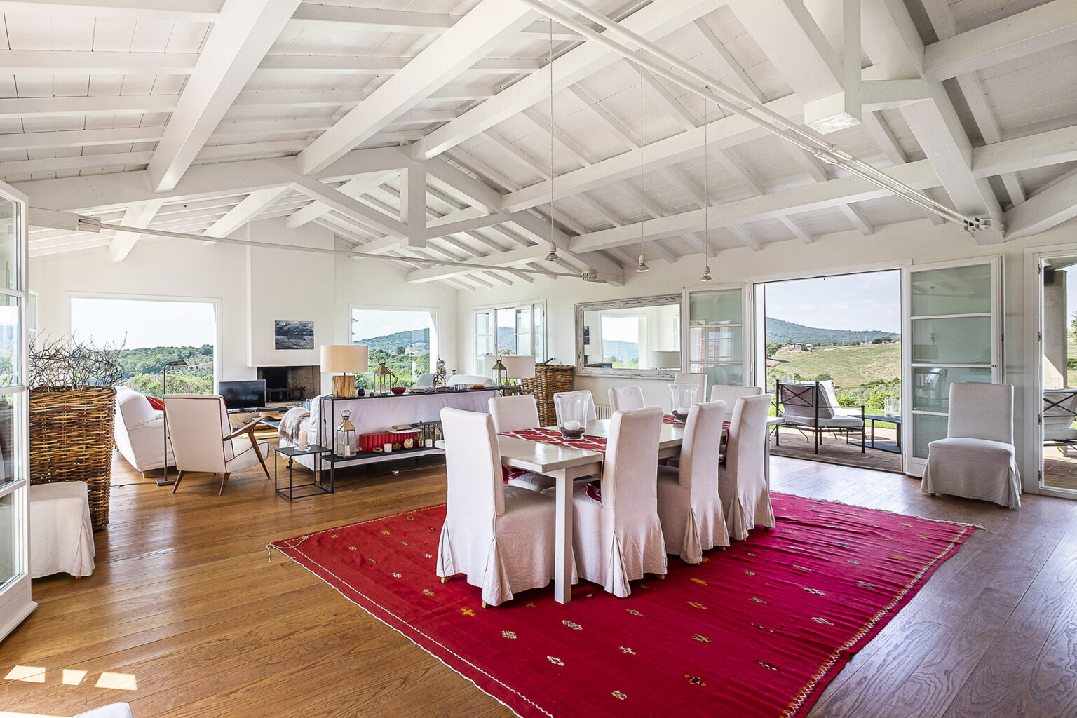 Bright and spacious living room with high white ceilings, wooden flooring, and large windows showcasing scenic views. The room features a dining area with a white table and upholstered chairs, a cozy seating area with modern furniture, and decorative elements like a red area rug and stylish lamps. | Only on SopranoVillas