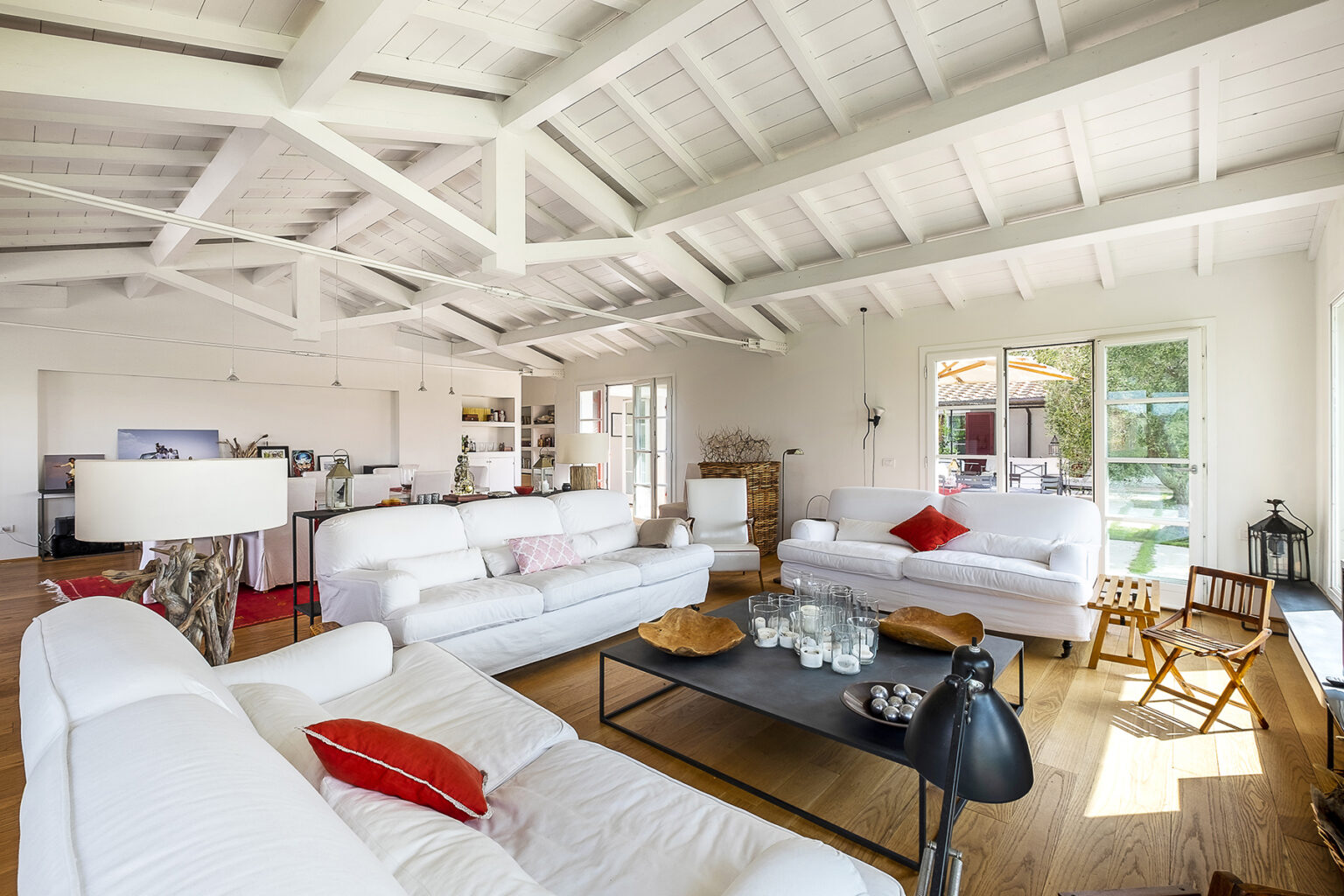 Bright and airy living room featuring white sofas with red accents, a wooden coffee table, and high wooden ceilings. Large windows provide natural light and a view of the outdoor patio, enhancing the spacious and modern aesthetic. | Only on SopranoVillas