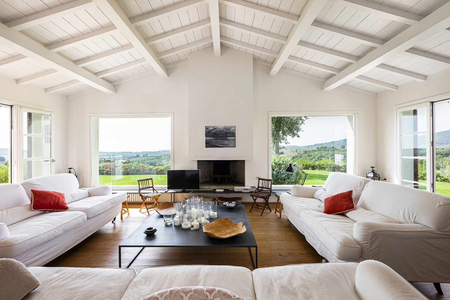 Spacious modern living room with white sofas, a black coffee table, and large windows offering panoramic views of the countryside. Features a minimalist design with wooden flooring, high ceilings, and a cozy fireplace, creating a bright and inviting atmosphere. | Only on SopranoVillas