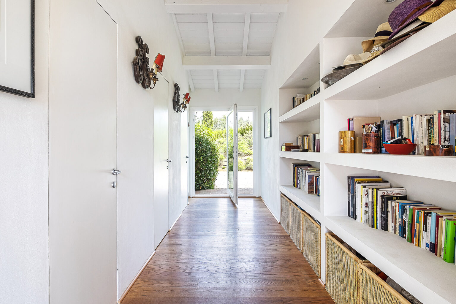 Bright and airy hallway featuring white walls and wooden flooring, with built-in shelves displaying books and decorative items, leading to an outdoor view. | Only on SopranoVillas