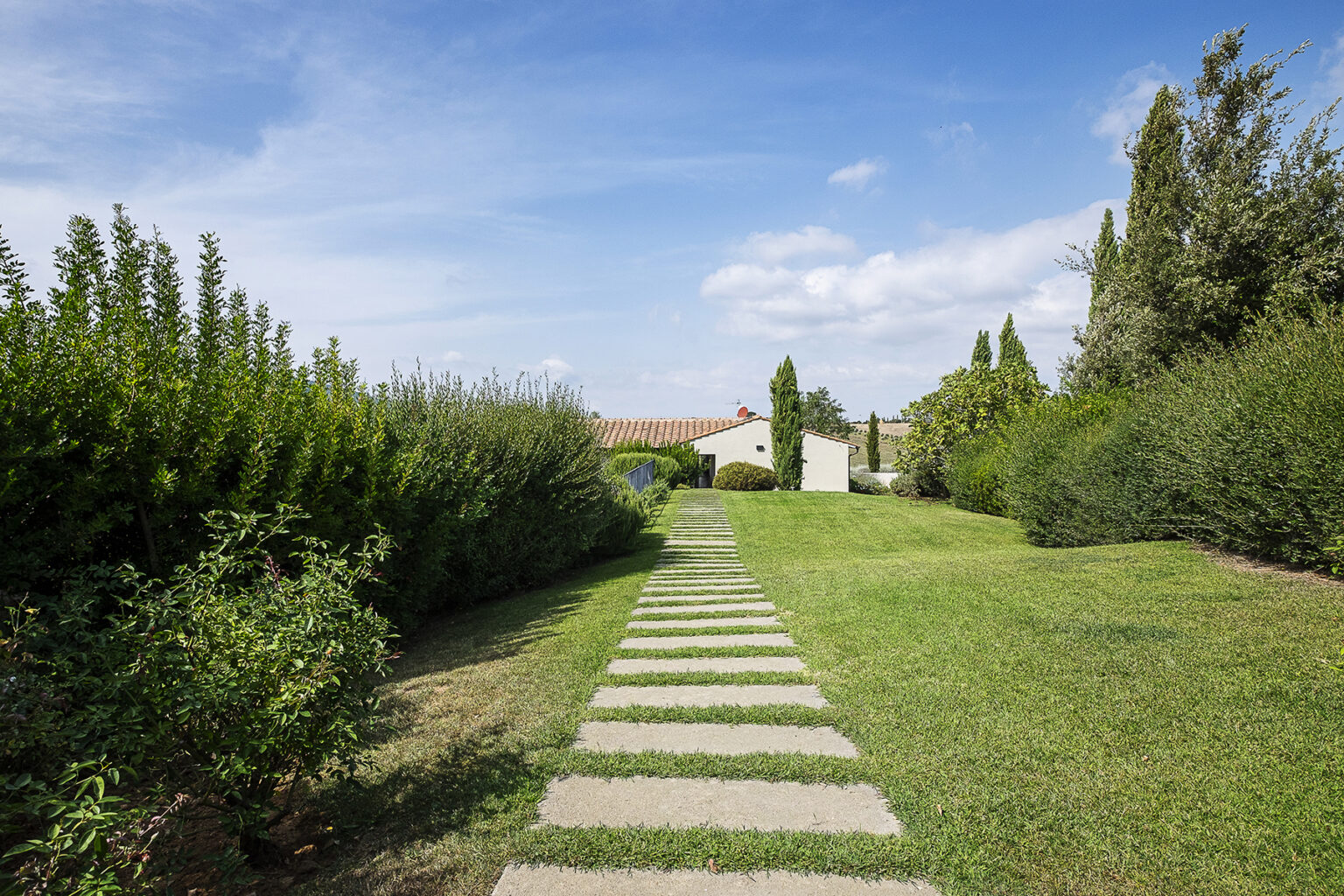 Pathway through a lush garden leading to a white building under a blue sky, surrounded by green shrubs and trees. Ideal for nature lovers and garden enthusiasts. | Only on SopranoVillas