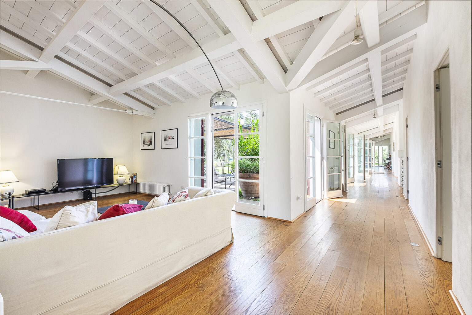 Spacious living room with high ceilings, wooden floors, and large windows, featuring a comfortable white sofa, a flat-screen TV, and an open hallway leading to an outdoor area. | Only on SopranoVillas
