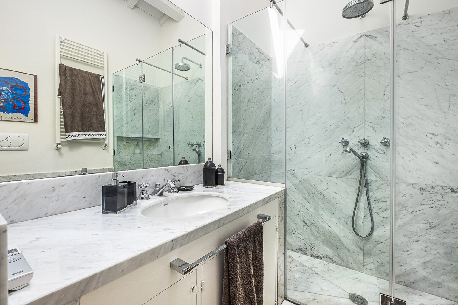 Modern bathroom featuring a marble countertop, glass shower enclosure, and contemporary fixtures. The design includes a sleek mirror and decorative artwork, creating a stylish and functional space. | Only on SopranoVillas
