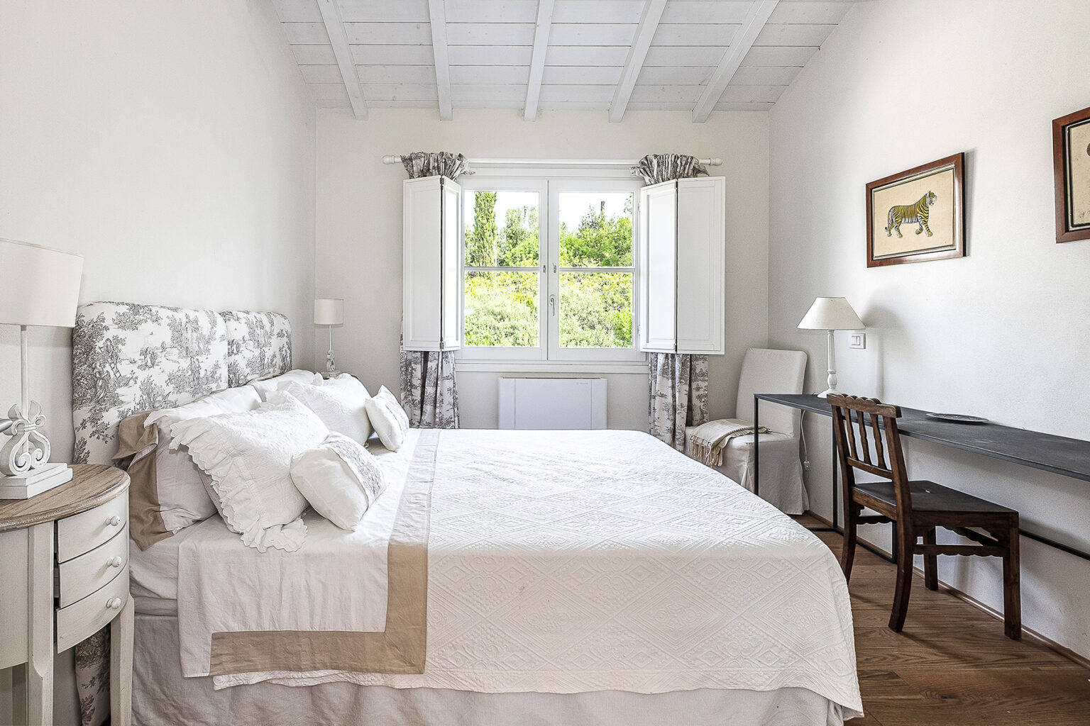 Bright and airy bedroom featuring a cozy double bed with decorative pillows, a wooden chair, and a desk, complemented by natural light from large windows with white curtains and a view of greenery outside. | Only on SopranoVillas
