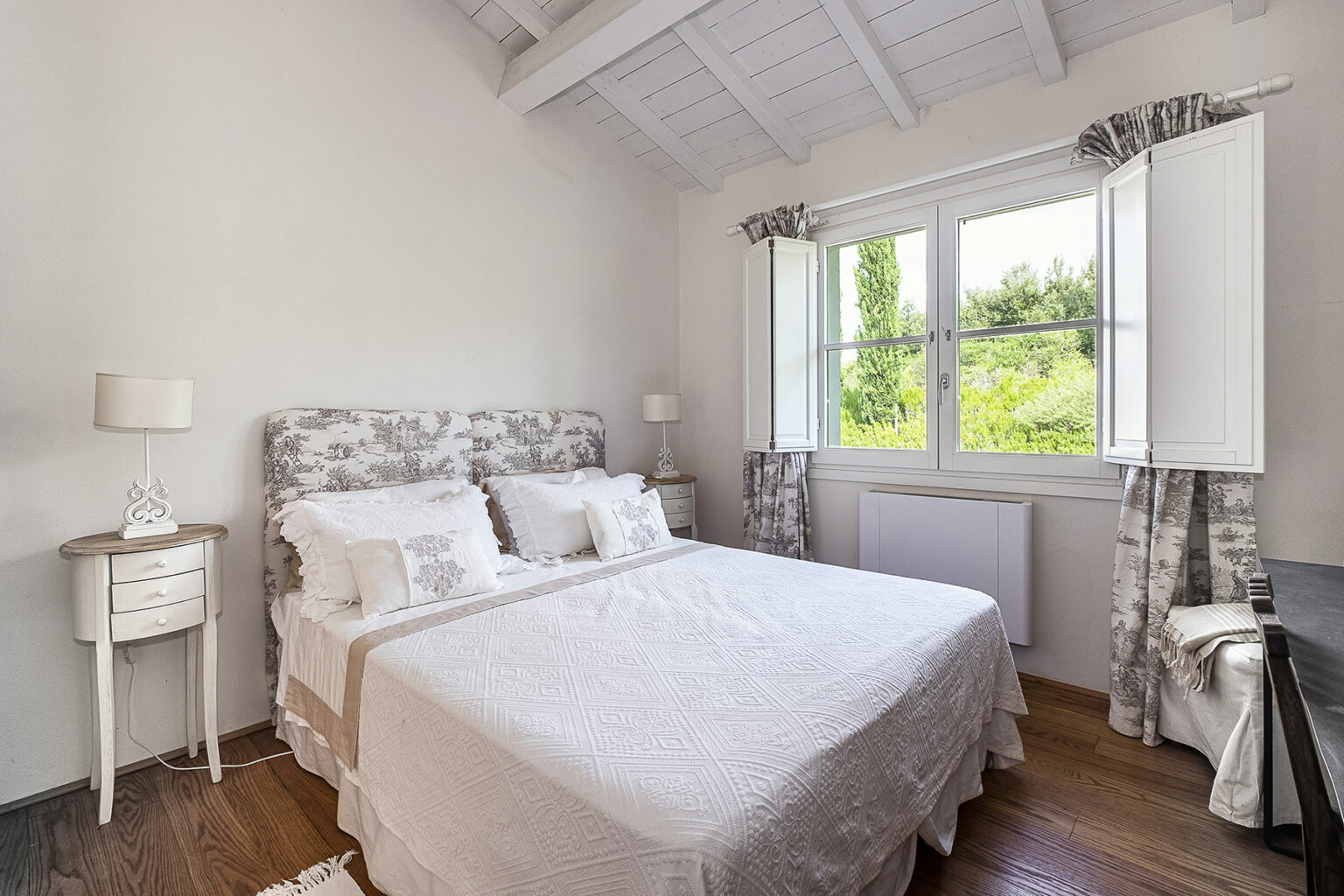 Bright and airy bedroom featuring a queen-sized bed with a floral headboard, white linens, and natural light streaming through open windows. The room includes a small bedside table and elegant curtains, complemented by wooden flooring and a cozy atmosphere. | Only on SopranoVillas