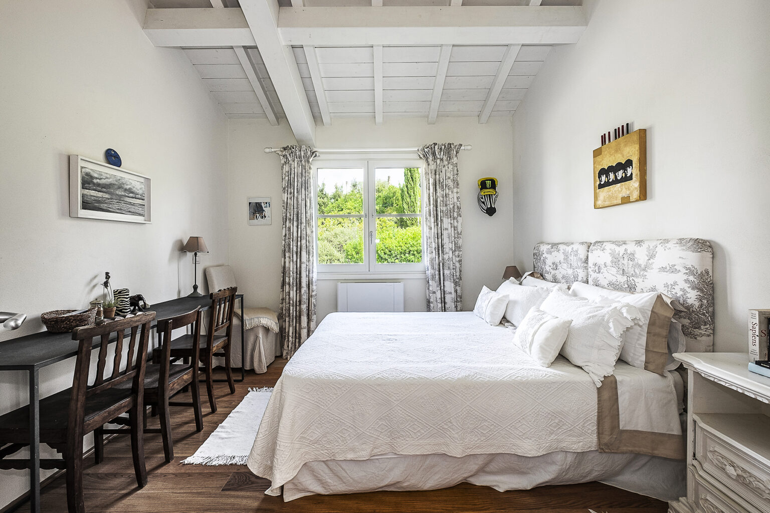 Bright and airy bedroom featuring two large beds with white linens, a wooden desk and chairs, and large windows with garden views. The room has a modern yet cozy design with decorative artwork on the walls and natural light flooding in. | Only on SopranoVillas
