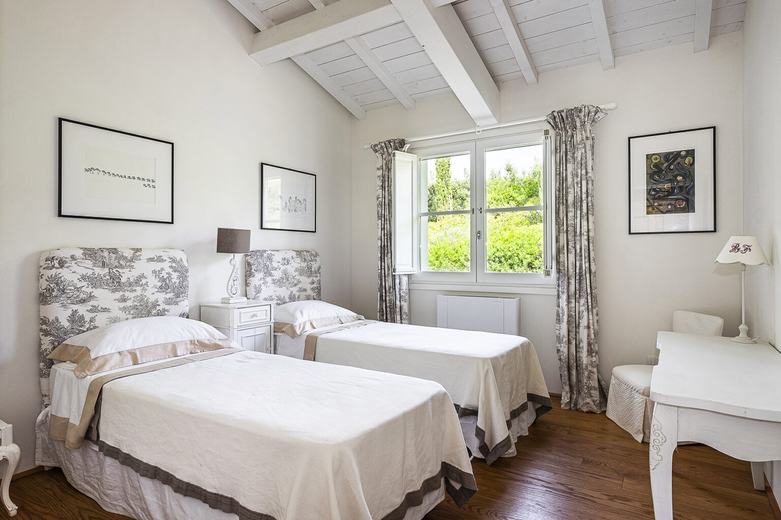 Bright and airy guest bedroom featuring two twin beds with floral headboards, large windows with natural light, and tasteful decor, including framed artwork and elegant curtains. Ideal for relaxation and comfort. | Only on SopranoVillas