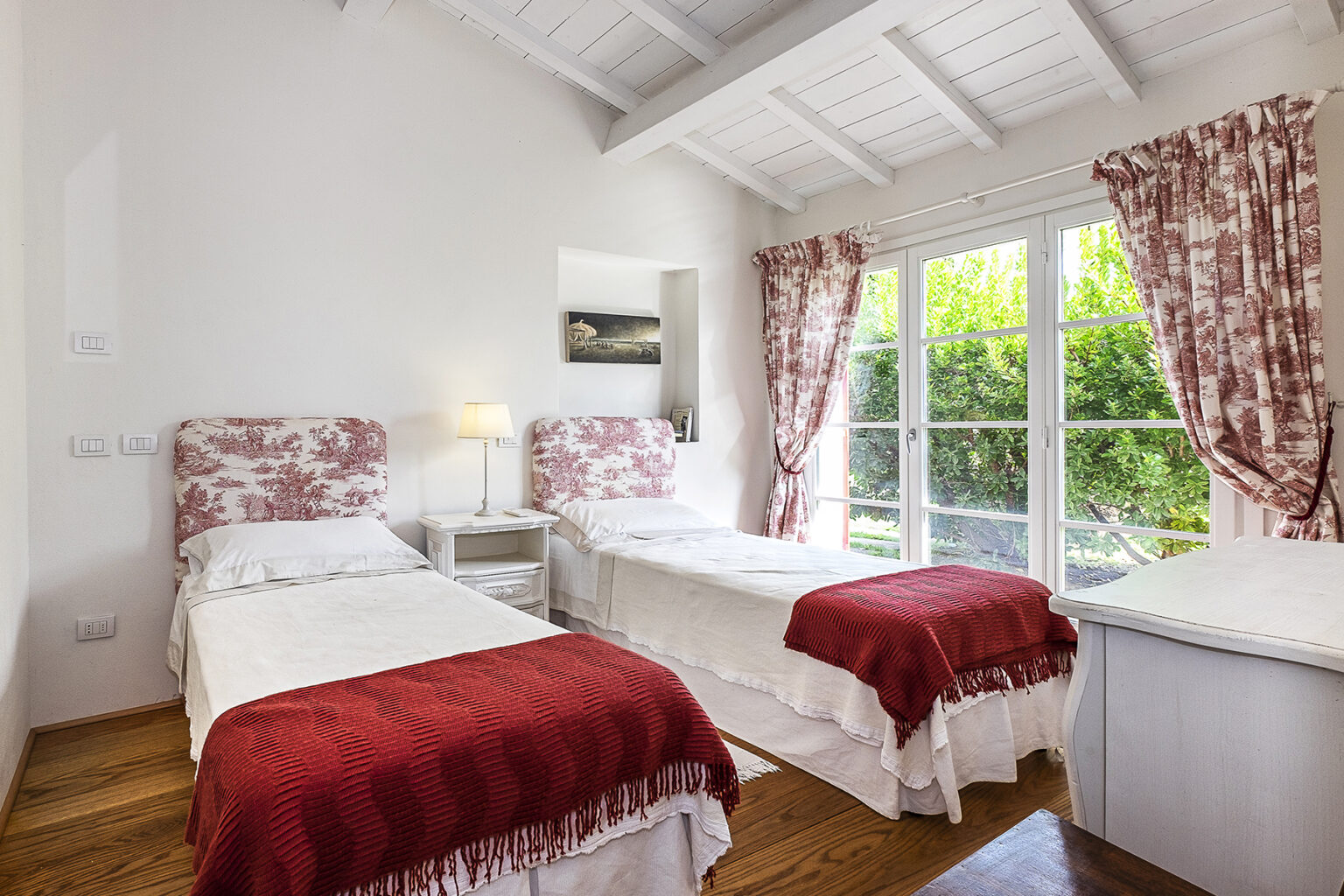 Cozy bedroom featuring two twin beds with floral headboards, red throws, and white linens. The room has natural light streaming through large windows, with elegant curtains and a bedside lamp. Perfect for a relaxing stay. | Only on SopranoVillas