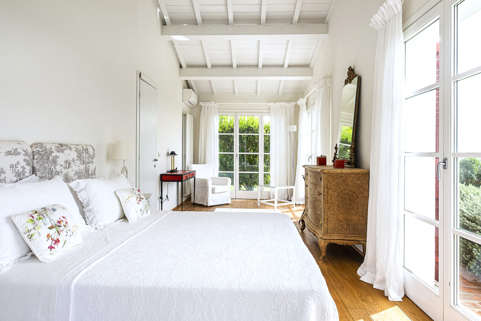 Bright and airy bedroom featuring a white quilted bedspread, floral accent pillows, and large windows with sheer curtains. The room includes a vintage wooden dresser, a modern armchair, and a stylish red side table, creating a cozy and inviting atmosphere. | Only on SopranoVillas