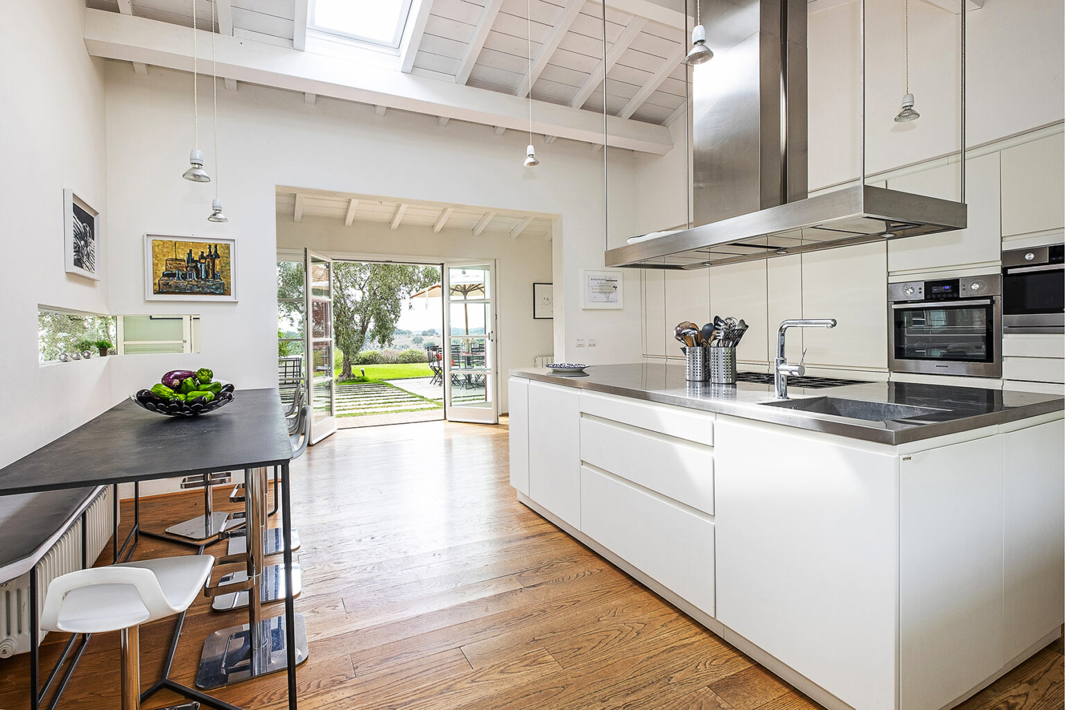 Modern kitchen interior featuring sleek white cabinetry, stainless steel appliances, and a wooden floor. Natural light floods the space through large windows, with a dining table set for two and a view of the garden outside. | Only on SopranoVillas
